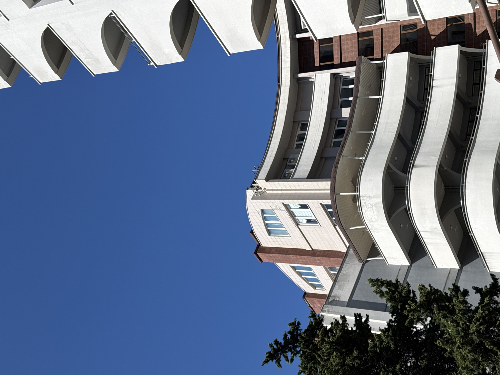 A low-angle, close-up shot captures the balconies of a modern, white, multi-story building against a clear, deep blue sky. The building features curved balconies with light gray railings, stacked in tiers. To the left, a portion of another building is visible, clad in light beige stone with red brick accents and white-framed windows. A dense, dark green coniferous tree partially obscures the left side of the image. The architecture suggests a contemporary residential or hotel complex.