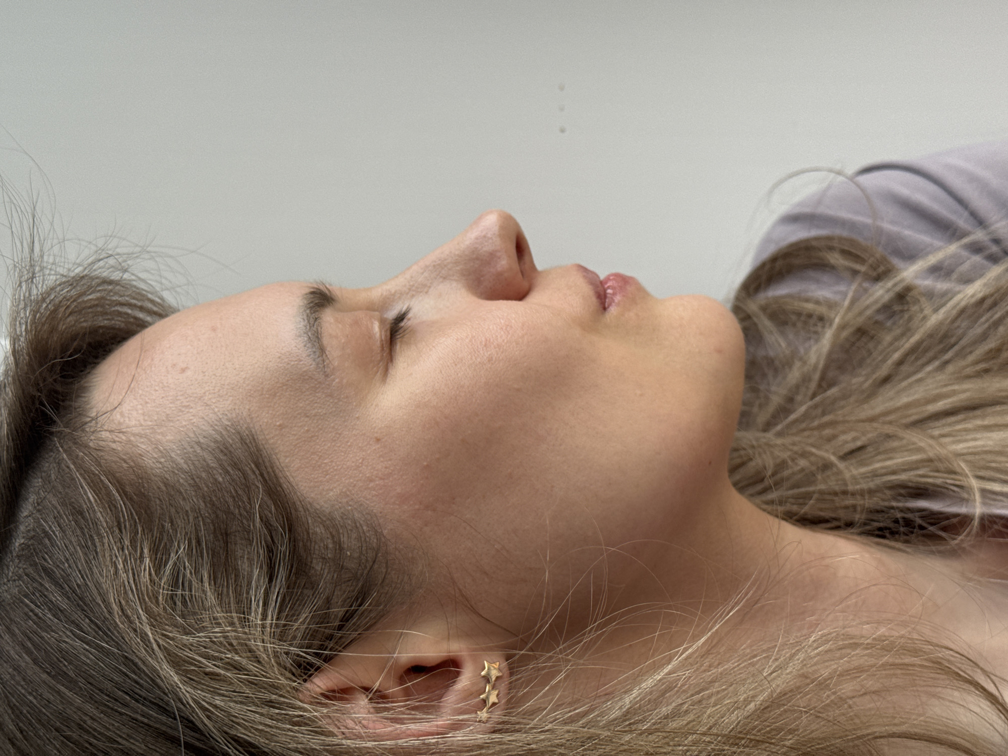 The image shows a person lying down with their face visible in profile. The person has light skin, closed eyes, and light brown hair with blonde highlights. They are wearing a small gold earring shaped like three stars. A portion of a light-colored garment is visible near the neck. The background appears to be a plain, light-colored wall. There is no information in the image to indicate its location or specific details about the city of Chaikovsky, Russia.