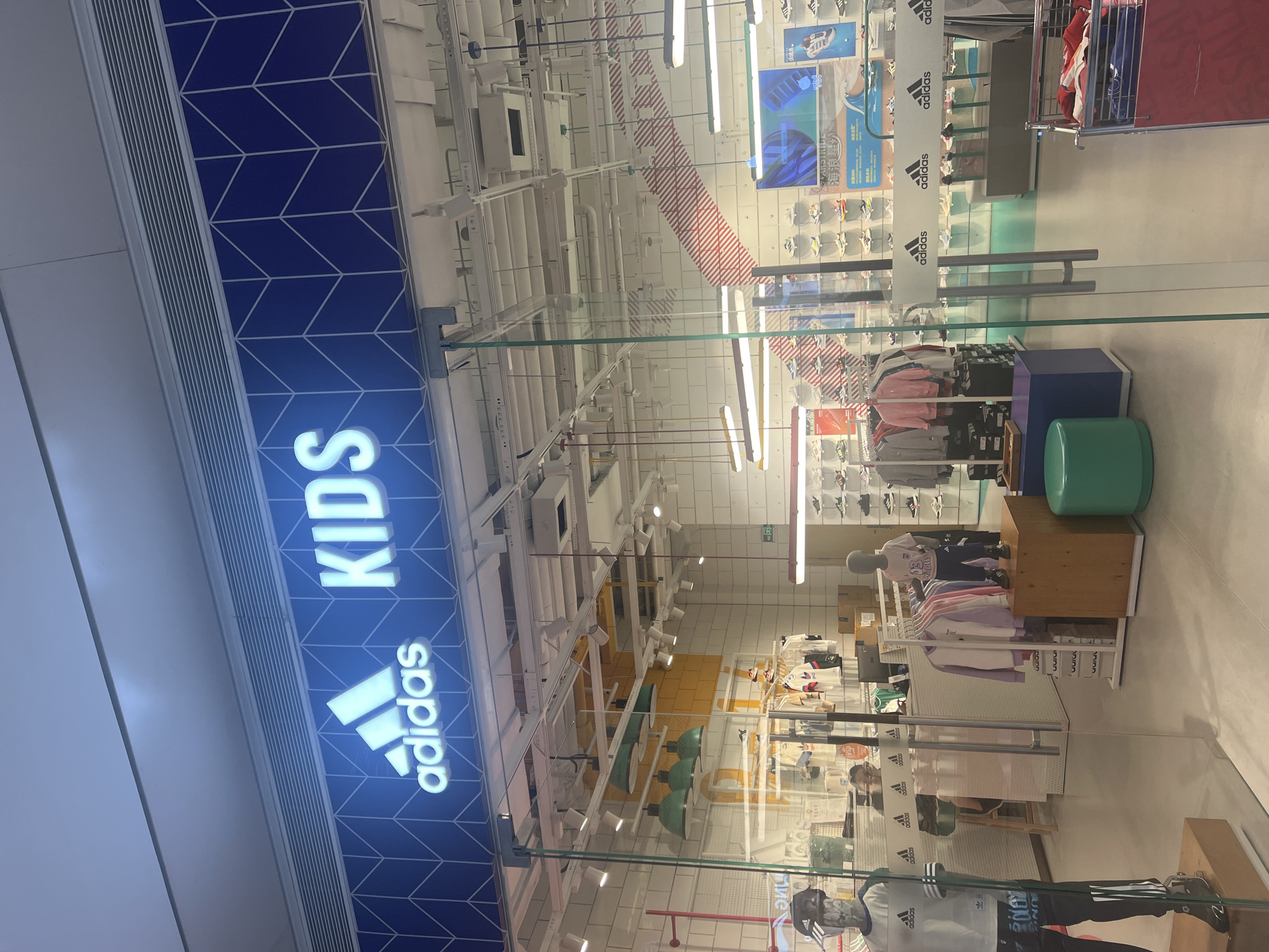 The image shows the exterior of an Adidas Kids store in Liandu District, China. The store features a large illuminated Adidas logo and the word "KIDS" in large white letters against a blue geometric background on the facade. Through the glass storefront, various Adidas merchandise is visible, including clothing on racks and mannequins, and shoes displayed on shelves. Several mannequins are dressed in Adidas clothing, and a child mannequin is on display inside. The store interior has white tiled walls, industrial lighting, and display shelves. Reflections of the interior are visible on the glass. The surrounding environment appears to be a shopping mall or commercial area, with other stores suggested by reflections.