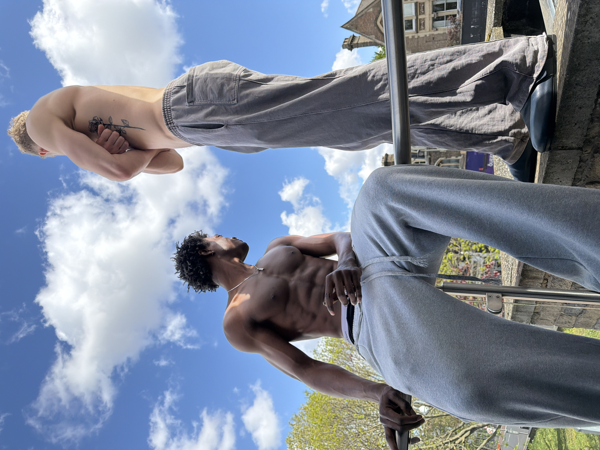 Two men are standing on a stone structure with metal railing. The man at the top is fair-skinned with a rose tattoo on his side and is wearing grey trousers and black shoes. The man below is dark-skinned with curly black hair and is wearing a silver necklace, grey sweatpants, and underwear. The background shows a bright blue sky with white clouds and a building with a spire. The image was likely taken in London, United Kingdom.