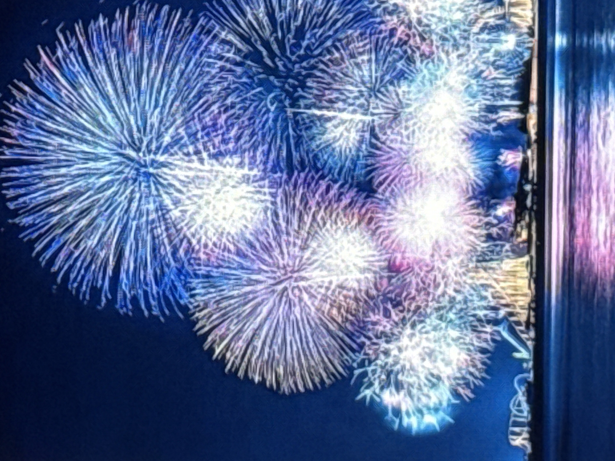 The image depicts a nighttime fireworks display over a city skyline, with the water in the foreground reflecting the colorful explosions. The fireworks are predominantly white, blue, and purple, with some pink and yellow hues. The city skyline in the background is dimly lit, with some structures appearing to be industrial or modern architecture. The water is dark and calm, with shimmering reflections of the fireworks. There are no people visible in the image. The setting is likely a coastal or riverside urban area at night, illuminated by the vibrant spectacle of the fireworks. The provided context about Norilsk, Russia, is not directly supported by visual evidence within the image itself.