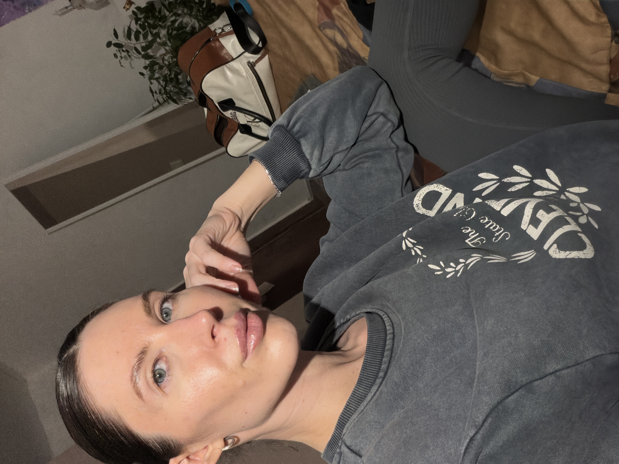 A young adult female is depicted in a portrait-style image, appearing to recline indoors. She has fair skin, light blue-green eyes, and dark hair pulled back from her face. Her expression is calm and direct as she looks towards the camera. She wears a natural makeup look, featuring glossy lips, and her left hand is positioned near her head. On her left wrist, a thin silver bracelet is visible, along with a silver hoop earring in her left ear.
She is dressed in a dark grey, possibly acid-wash or faded, crewneck sweatshirt. The sweatshirt features white graphic elements including leaf motifs and partial text such as "IEVND" and "The State Of...". Below the sweatshirt, she appears to be wearing dark grey leggings or similar casual bottoms.
The background suggests an indoor setting, possibly a room or bedroom. In the upper left, a brown and off-white shoulder bag with black straps is partially visible, with text resembling "SVSBAG" on its side. Behind the bag, a green potted plant adds a natural element. A light brown or beige textured fabric, possibly part of bedding or furniture, occupies the right side of the background. The wall or ceiling is plain and light-colored.
The image is taken from a low angle, with the subject positioned on her side due to the rotated perspective. The lighting is soft and ambient, suggesting an indoor environment with artificial light or low natural light, possibly in the evening. There are no specific visual cues within the image to identify the location, but the provided context states it is Perm, Russia.
