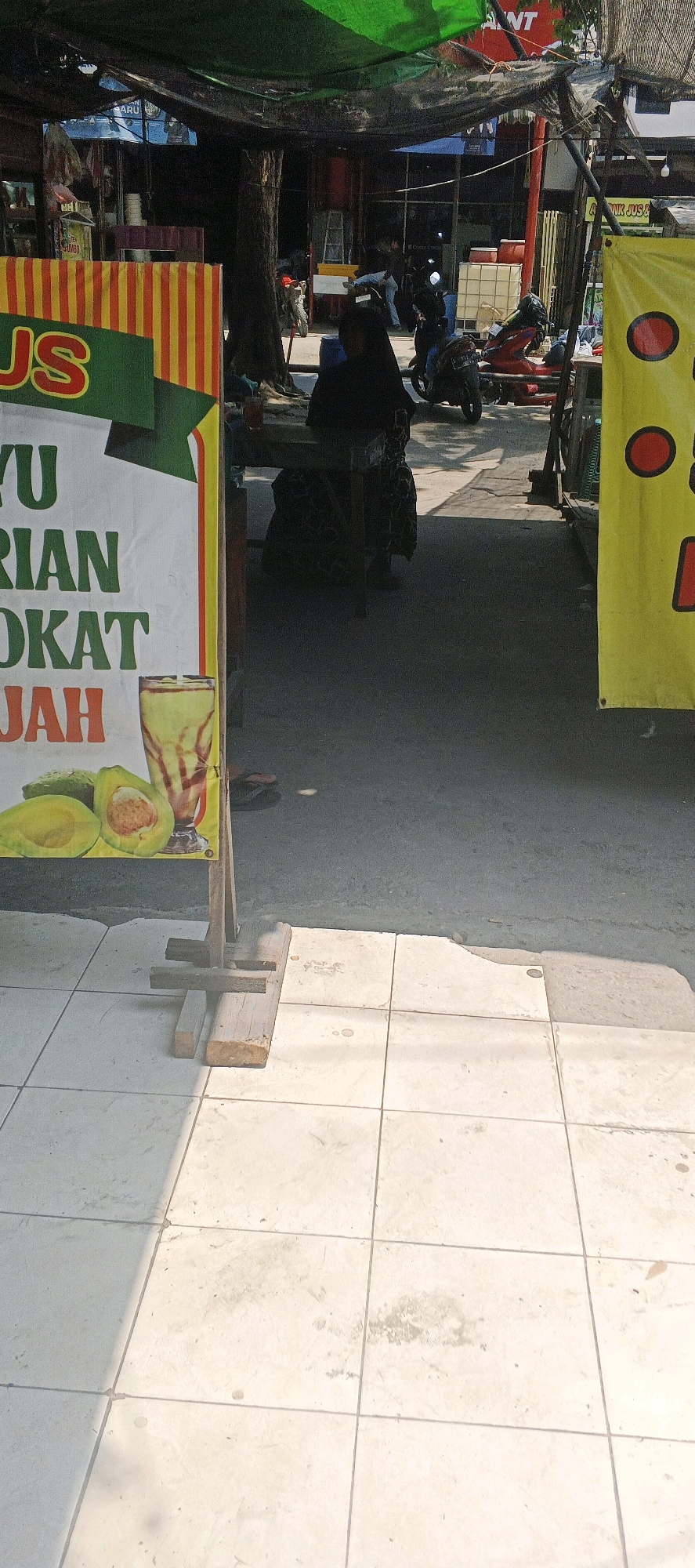 A street scene in Maduran, Indonesia, features a stall with a banner advertising "JUS YU RIAN OKAT JAH". The banner, adorned with red and yellow vertical stripes, displays the text in green and orange. In front of the banner, a glass of avocado juice sits on a table, alongside a cut avocado. A person wearing a black hijab and traditional attire is seated at the table. The area is paved with light-colored square tiles, with a portion of asphalt visible in the background. The stall is covered by a green tarp. Further back, several motorcycles are parked, and other individuals can be seen. A yellow banner with red circles is visible on the right side of the image. The lighting suggests it is daytime, with shadows cast by the sun.