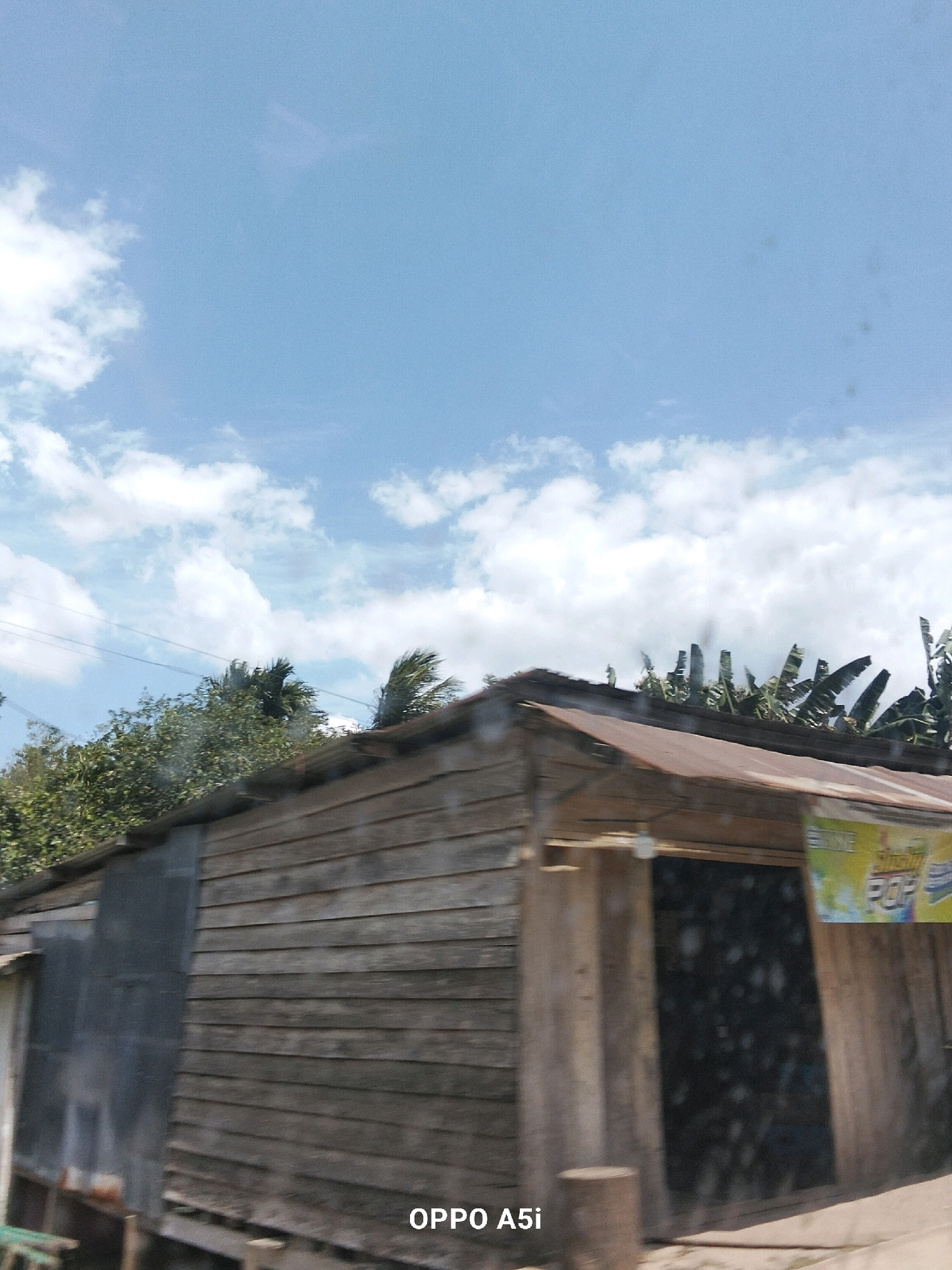 A rustic, weathered wooden building with a corrugated metal roof stands in the foreground of this outdoor, daytime photograph. The building appears to be a shop, with a dark, boarded-up window and a colorful sign advertising "POP" on the right side. Behind the building, lush green trees and banana plants are visible, swaying slightly in what appears to be a gentle breeze. The sky above is a bright blue, dotted with fluffy white clouds, suggesting fair weather. The overall scene evokes a simple, rural atmosphere, possibly in a tropical region. The timestamp "OPPO A5i" is visible in the lower right corner, indicating the device used to capture the image. The location of Puusangi, Indonesia is not evident from the image itself.