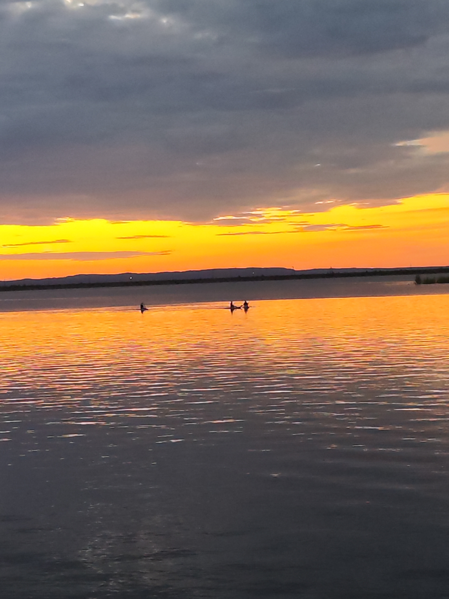 A sunset scene over a body of water in Chaikovsky, Russia, features a dramatic sky transitioning from a bright yellow and orange band near the horizon to dark, cloudy grey overhead. The water reflects the vibrant colors of the sky, creating a mirror-like surface with subtle ripples. On the water, two small human figures are visible, each appearing to be in a kayak or a similar small watercraft. They are positioned in the middle ground, spaced apart from each other. The distant shoreline is a dark, hazy silhouette of hills or landforms.