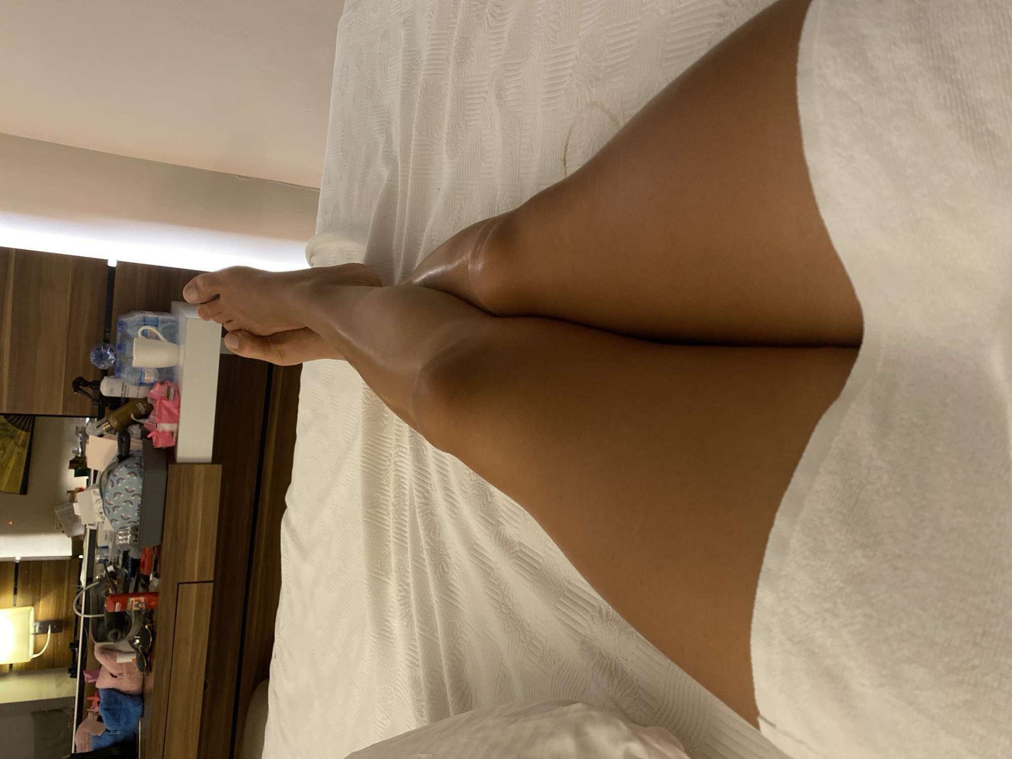 A person's legs, from the knees down, are shown crossed and resting on a white textured bedspread. The person's feet are bare and visible at the top of the frame. A white towel is draped over the lower portion of the legs. In the background, a wooden dresser is visible, holding various items including makeup bags, bottles, and a mug. A lamp is mounted on the wall above the dresser. The scene appears to be indoors, likely in a hotel room. The location is not specified in the image.