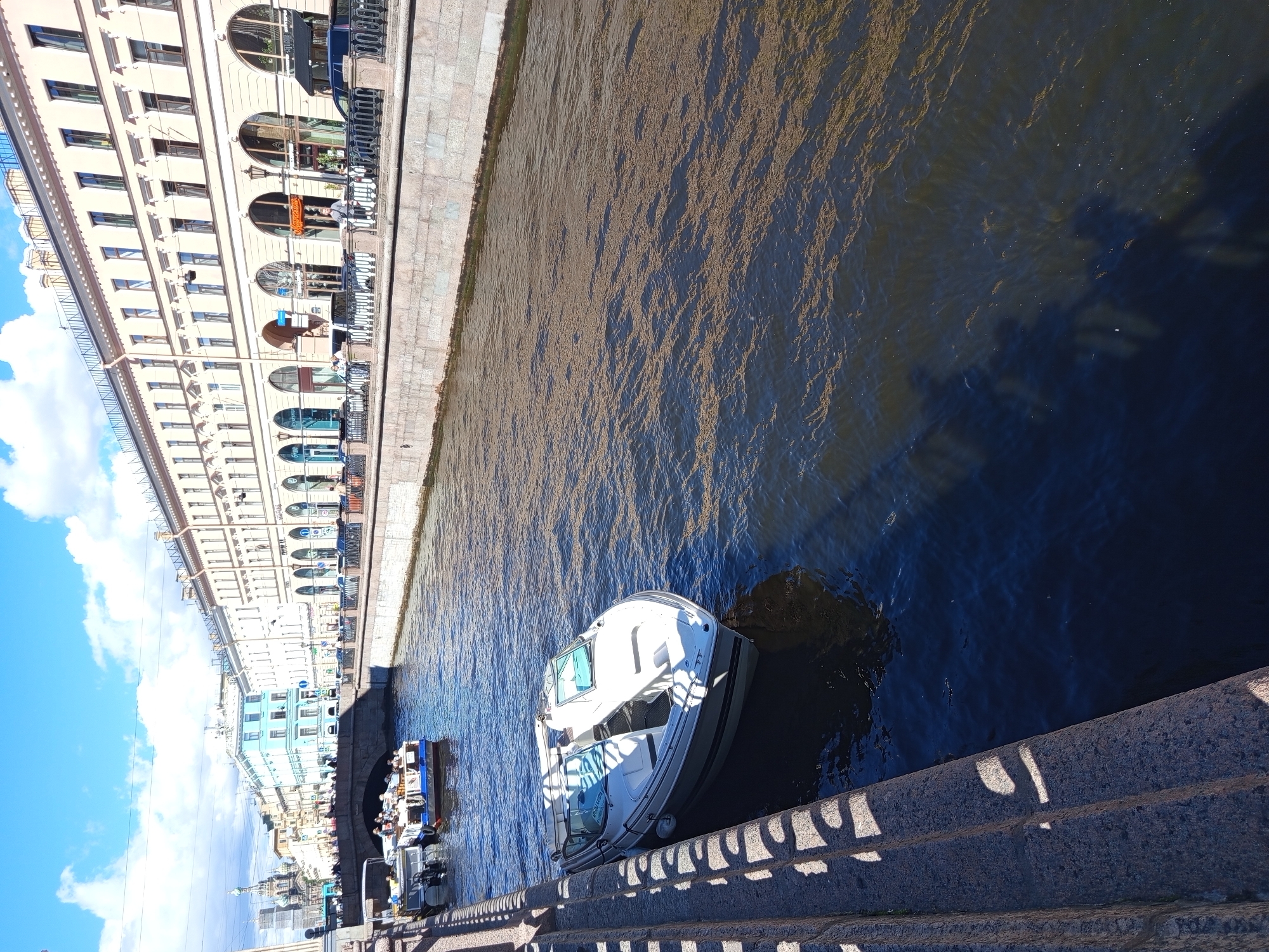 The image depicts a sunny day in Saint Petersburg, Russia, along a canal. In the foreground, a white motorboat is moored to a stone embankment. The boat has a gray stripe along its waterline and is equipped with two windshields. Further down the canal, two other boats are visible, one appearing to be a tour boat with passengers. The canal is lined with stone embankments and buildings with classical architecture. The buildings feature multiple windows, arched doorways, and decorative cornices. A pedestrian walkway with a black metal railing runs along the embankment in front of the buildings. People can be seen walking on the walkway and seated at outdoor cafes. In the background, a bridge spans the canal, and beyond that, a building with onion domes, likely a church, is visible against a blue sky with white clouds. The water in the canal is dark and reflects the sunlight, with some debris or floating particles visible on the surface. Shadows from the railing and the embankment are cast onto the stone steps in the left foreground.