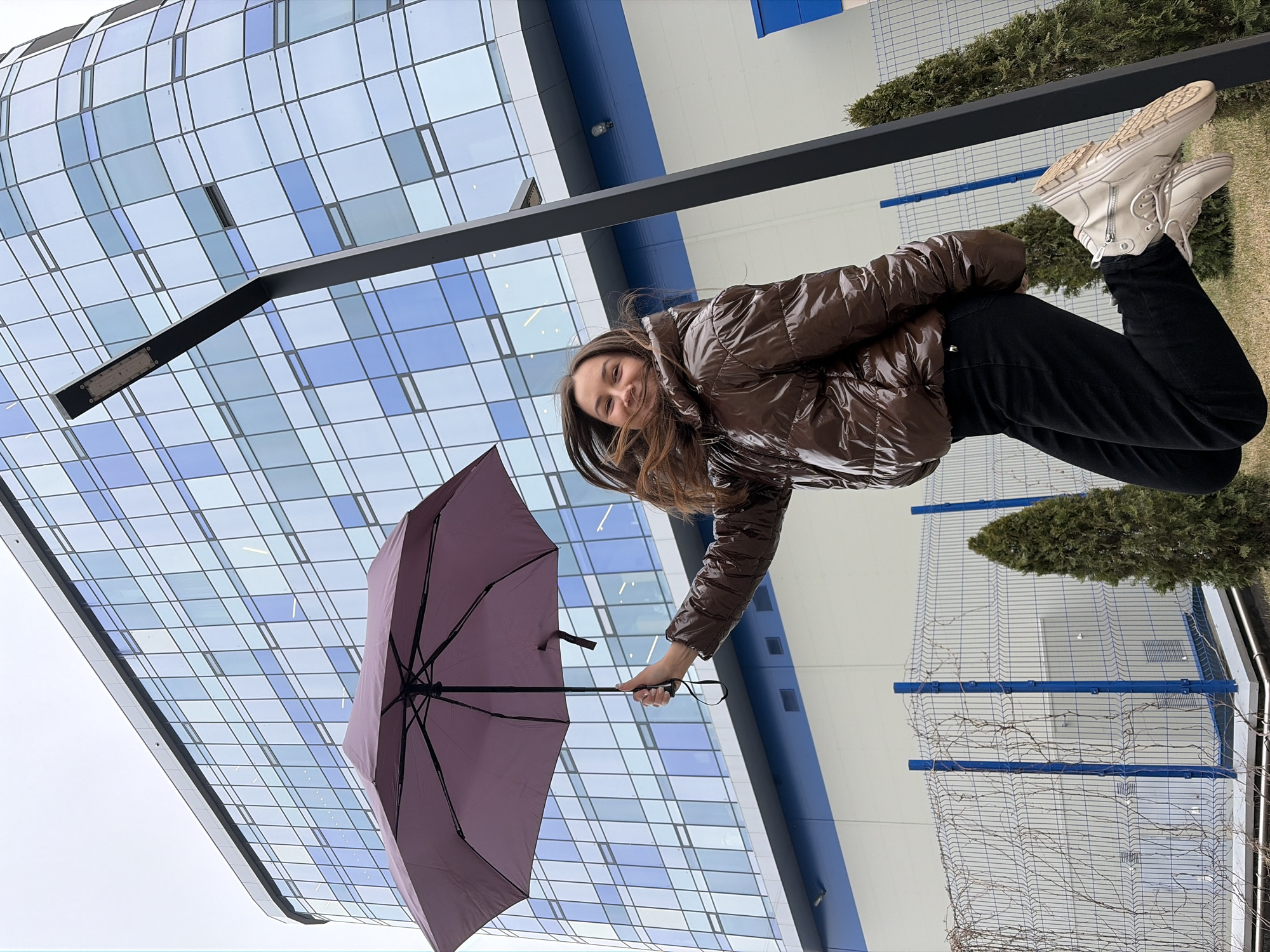A woman stands on grass near a blue-and-white building in Izhevsk, Russia, holding an open purple umbrella. She is wearing a shiny brown puffer jacket, black pants, and white boots. A black horizontal metal bar with a light fixture is positioned above her. A blue metal fence and small evergreen trees are visible in the background. The building's facade is constructed from light blue and white panels, creating a grid-like pattern.