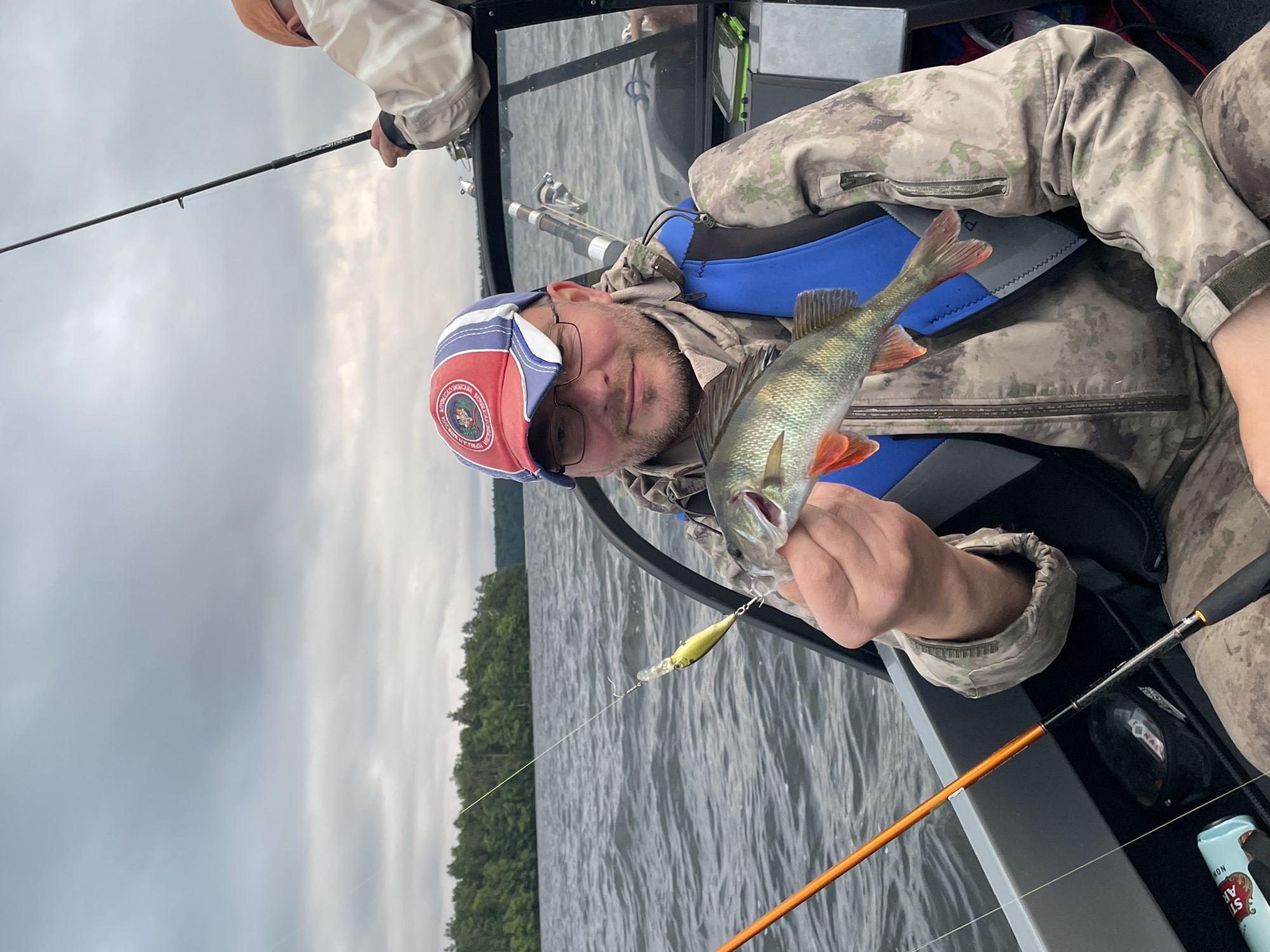 A middle-aged Caucasian man wearing glasses, a camouflage jacket, a blue life vest, and a red, white, and blue baseball cap is seated in a boat, holding a small perch he has caught. The man is looking towards the camera with a slight smile. Behind him, another person in a light-colored jacket and an orange cap is also in the boat, holding a fishing rod. The boat is on a body of water with choppy waves, and a treeline is visible in the distance under a cloudy sky. The scene appears to be in Yablonovka, Russia.