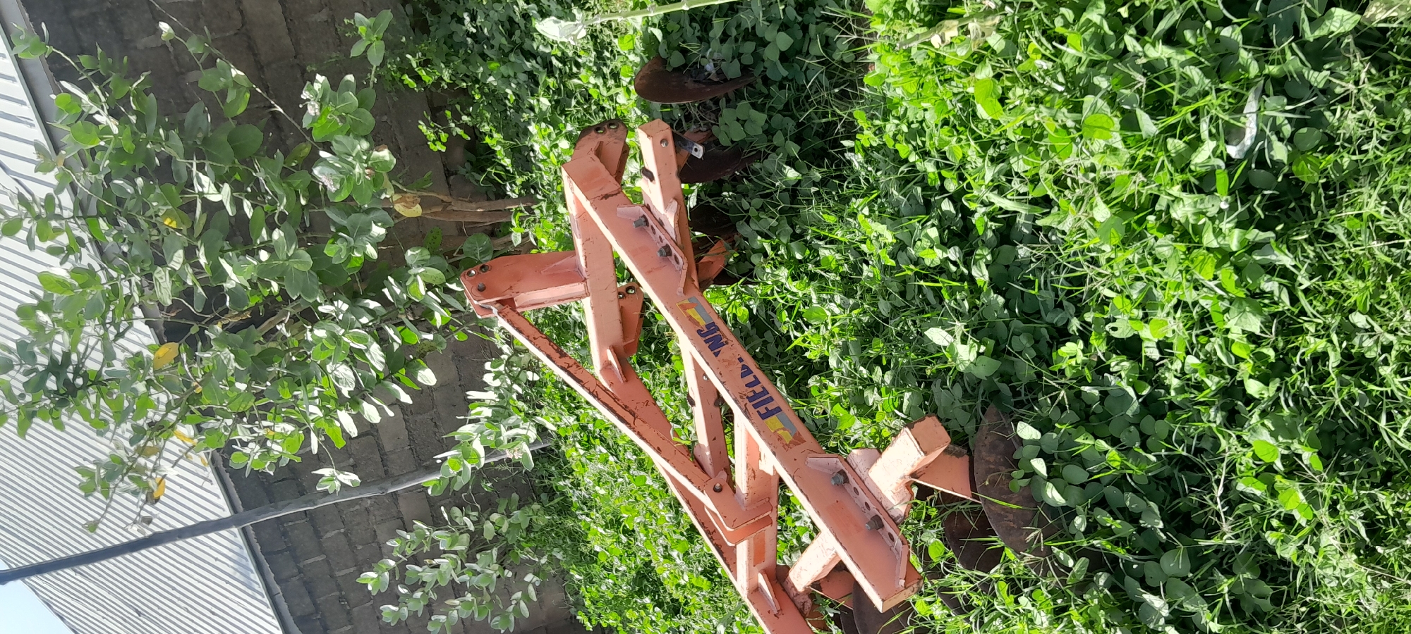 The image shows a piece of agricultural equipment, likely a plow or disc harrow, resting on a grassy slope. The equipment is orange and appears to be made of metal. There are multiple discs visible, suggesting it is a multi-disc plow. The background consists of lush green vegetation and a brick wall. Above the wall, a section of a corrugated metal roof is visible. The scene is outdoors, with sunlight illuminating the area. The text "FIELDING" is visible on the side of the equipment, indicating the manufacturer or brand. The context of Jalingo, Nigeria, suggests this scene might be in a rural or semi-rural area on the outskirts of the city, where agricultural activities are common. There are no people or animals visible in the image.