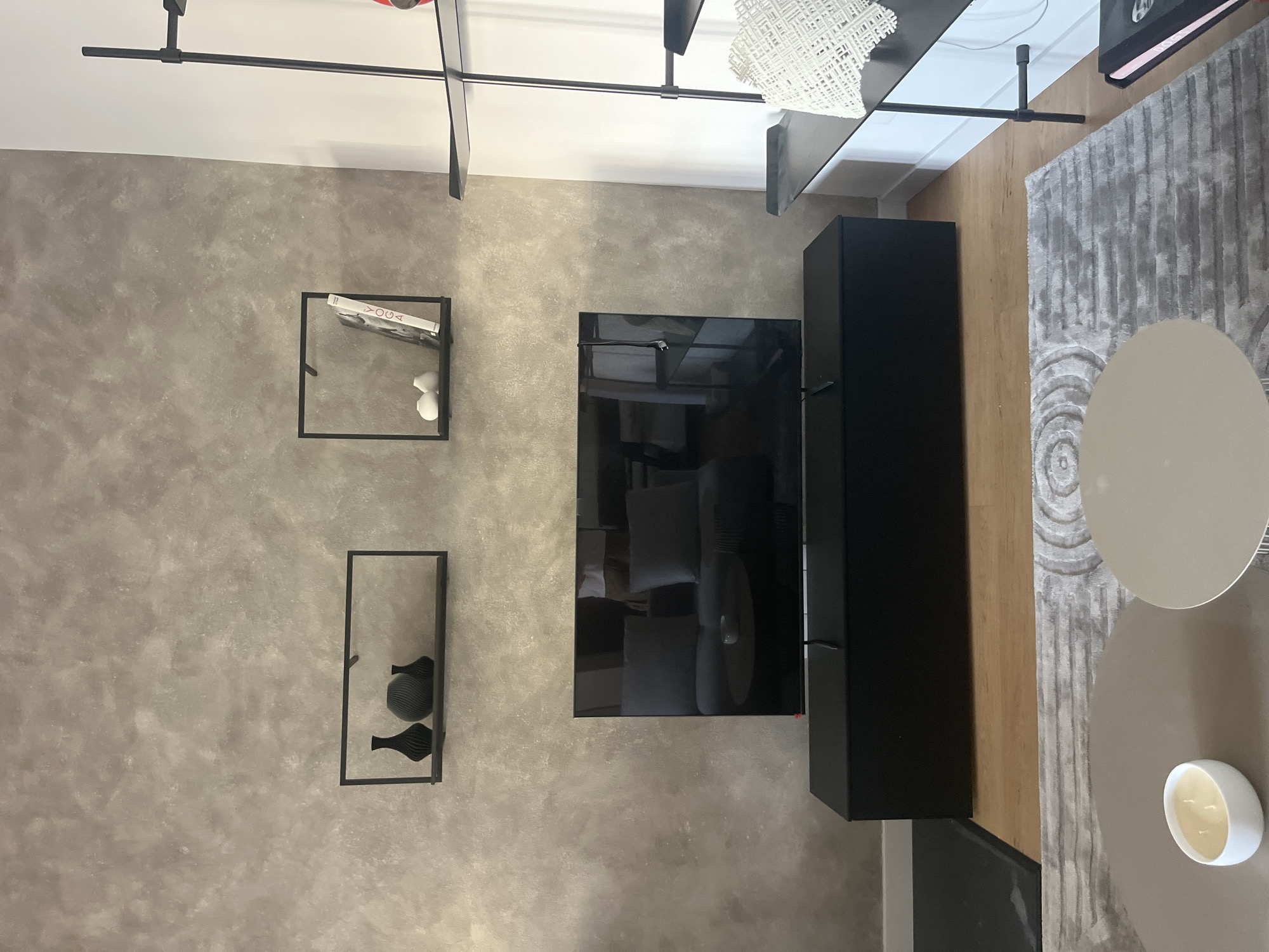 The image depicts a modern living room with a grey, textured accent wall. Mounted on this wall are two black, minimalist shelves. The left shelf holds three black vases of varying sizes and shapes. The right shelf displays two small white decorative spheres and a book titled "YOGA" in white lettering against a black cover.

Below the shelves, a large, flat-screen television is centrally positioned on a sleek black media console. The television screen reflects a dark gray sofa with two dark gray cushions and a small round coffee table with a light gray object on it.

To the right of the television setup, a tall, black multi-tiered shelving unit is visible against a white wall. The upper shelves of this unit are empty, while the lower shelves are partially visible, holding a white decorative object with a woven pattern.

In the foreground, a gray, patterned rug covers a portion of the wooden floor. Two round coffee tables are placed on the rug. The left coffee table has a white, oval-shaped candle with three wicks. A portion of a black book with a pink spine is visible on the floor to the right of the shelving unit. The scene appears to be an interior residential space, likely a living room. No people are present in the image.