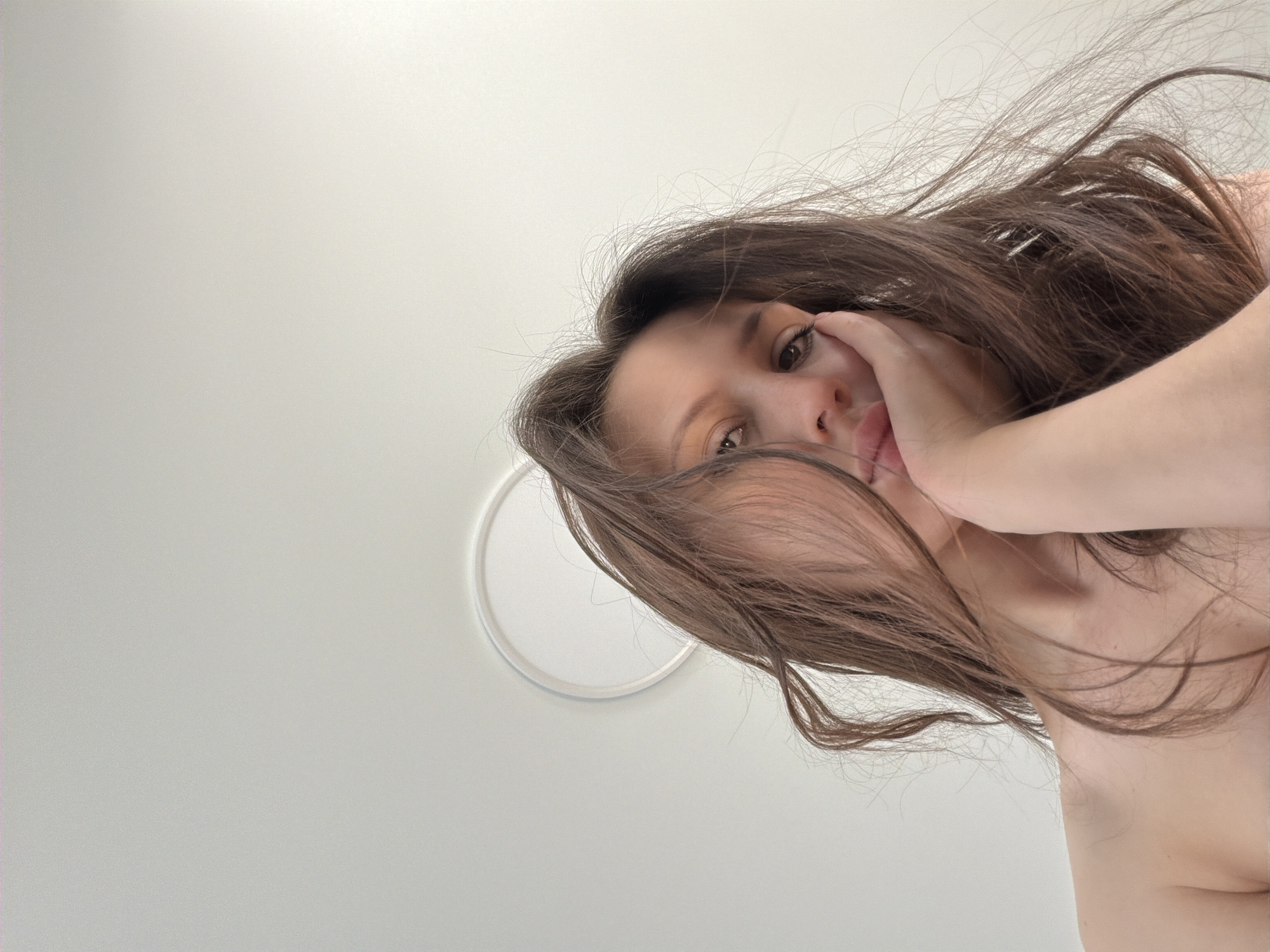 The media file is a low-angle, close-up, outdoor selfie of a young woman. She has long, dark brown hair that is blowing across her face, obscuring part of her features. Her right hand is resting on her cheek and near her mouth. She is looking towards the camera with a neutral expression. Her skin appears fair, and her eyes are brown. She is not wearing any visible clothing on her upper body.
The background is a plain, light-colored surface, possibly a wall or ceiling, with a circular white fixture visible to the left of the woman's head. The lighting suggests it is daytime, with diffused light that is not harsh. There are no discernible weather cues.
The image appears to have been taken in a residential setting, likely indoors given the clean, uniform background and the ceiling fixture. The overall mood of the image is casual and intimate. No text is visible in the media file. The location context is Khimki, Russia.