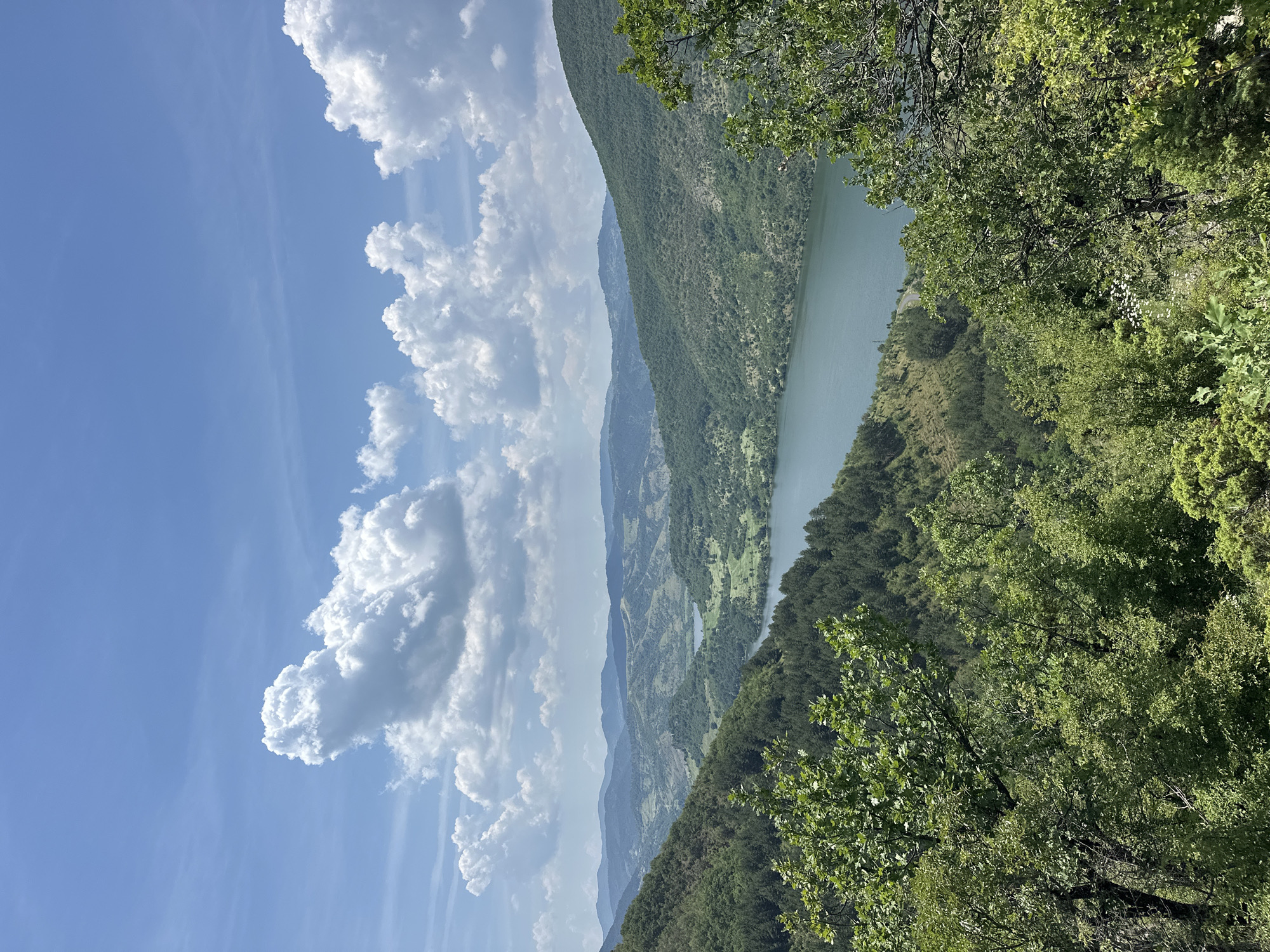 The image displays a panoramic landscape scene captured from an elevated viewpoint, depicting a natural environment in or near Ilinitsa, Bulgaria. The upper portion of the frame features a bright blue sky dotted with scattered, fluffy white cumulus clouds, indicating fair weather conditions. Below the sky, a vast expanse of rolling hills and mountains dominates the middle ground and background, covered extensively with dense, vibrant green foliage, primarily trees.
A prominent body of water, appearing as a lake or a wide river, meanders through the central valley, its surface reflecting a deep greenish-blue hue. Further into the distance, a second, smaller body of water is visible nestled among the hills. The landscape exhibits varied topography, with forested slopes giving way to gentler gradients in the distance, potentially indicating agricultural patches or clearings. The foreground consists of the tops of lush green trees and shrubs, framing the view and suggesting the vantage point is within a forested area. No individuals, man-made structures of significant scale, or discernible actions are visible within the frame. The overall impression is one of a tranquil, expansive natural landscape during daylight hours.