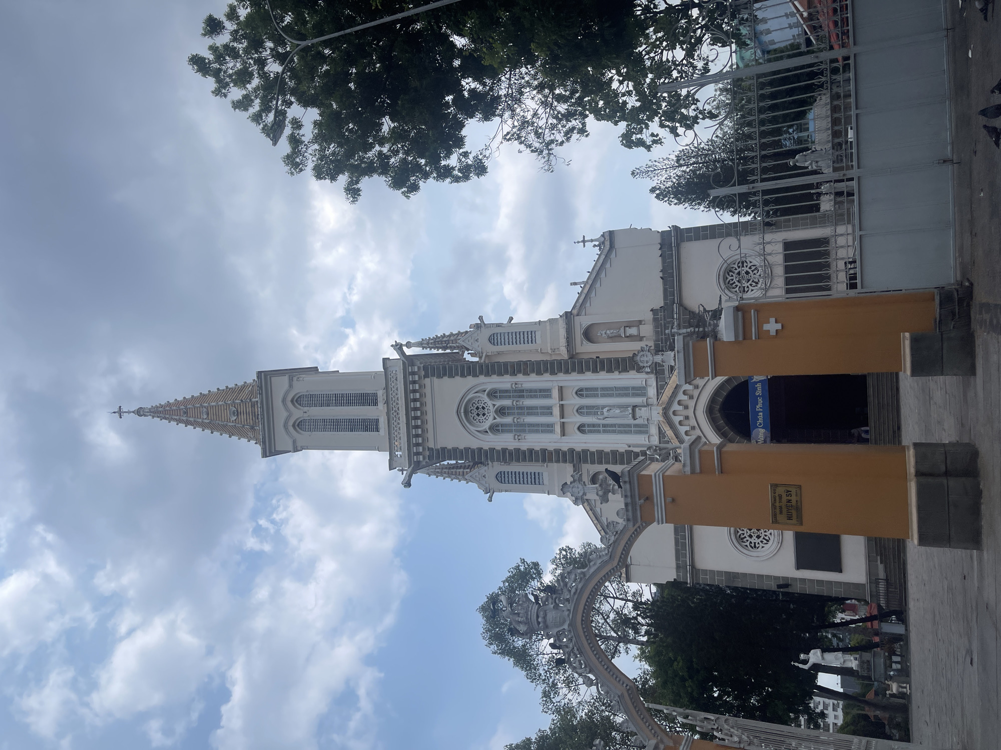 The image depicts the Thủ Đức Church in Vietnam. The church is a tall, white building with a spire. The sky is cloudy with some patches of blue. Trees are visible in the foreground and background. A metal gate is visible on the right side of the frame. The entrance to the church has yellow pillars on either side. A sign written in Vietnamese is visible. People are visible inside the church. The image is taken from a low angle.