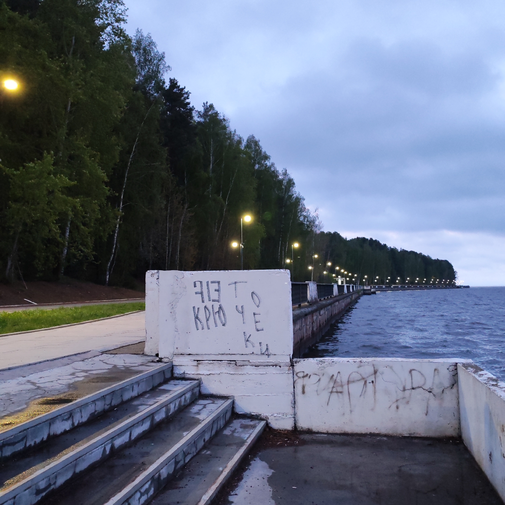 The image depicts a concrete pier or embankment along a body of water, likely the Kama River, in Chaikovsky, Russia. The sky is overcast. The pier has streetlights extending into the distance. The concrete structure in the foreground has graffiti on it. There are stairs leading down to the lower level. A forested area lines the land adjacent to the pier. The water shows signs of waves or ripples, indicating some wind activity.