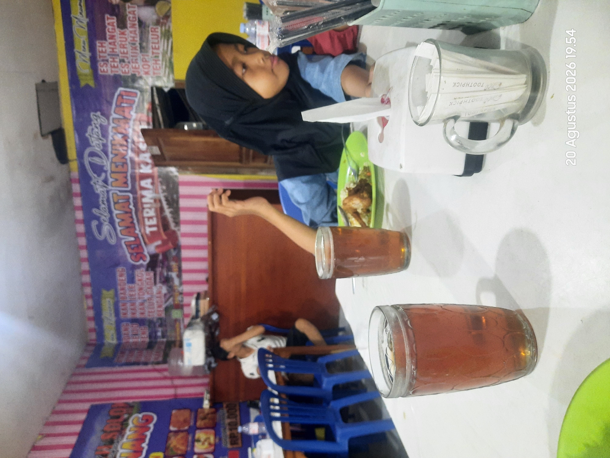 A young girl wearing a black hijab and blue shirt is seated at a white table. She is looking down at a plate of food. In front of her are two glasses of tea and a tissue box. To her left, a man in a white shirt is seated at another table. Blue chairs are visible in the background. The walls are decorated with pink and white stripes. A banner in the background reads "Selamat Datang". The timestamp "20 Agustus 2026 19.54" is superimposed on the image. The scene appears to be a casual dining establishment.