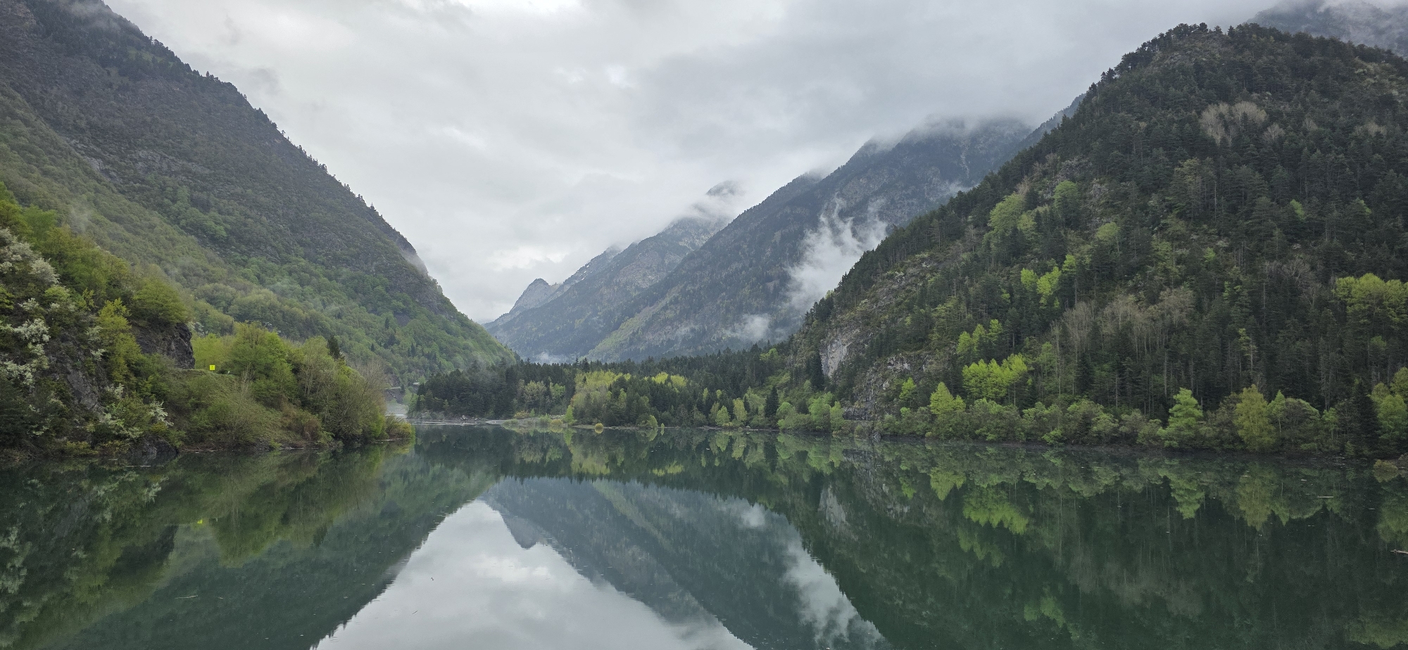 A panoramic landscape photograph captures a serene mountain lake or wide river nestled in a valley surrounded by steep, densely forested mountains. The sky is overcast, with low-hanging clouds and mist clinging to the upper slopes and peaks of the mountains, suggesting a cool, damp atmosphere.
The dominant subjects are the majestic mountains, which rise sharply from the water's edge, and the lush vegetation covering them. The trees display a variety of green hues, from the dark, deep green of coniferous forests to lighter, brighter greens of deciduous trees, possibly indicating spring or early summer.
The water surface is remarkably still, creating a nearly perfect, mirror-like reflection of the mountains, trees, and cloudy sky. This reflection enhances the depth and symmetry of the scene. On the lower slopes of the mountain to the left, a small, bright yellow rectangular sign or marker is visible amidst the foliage, reflecting faintly in the water below. No other human structures or activities are discernible.
The overall impression is one of tranquil natural grandeur, characteristic of a high-altitude mountain region like Benasque, Spain, with the misty conditions adding a sense of peaceful solitude and dramatic atmosphere. No legible text is visible in the image.