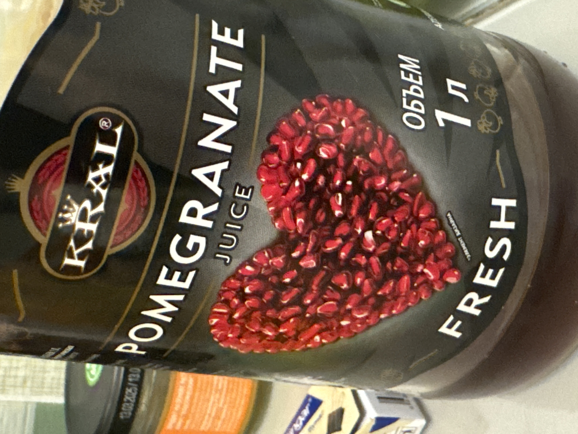 The image presents a close-up, slightly tilted view of a bottle of "KRAL" Pomegranate Juice. The bottle is dark brown or black, and its label, predominantly black with white and gold lettering, is clearly visible.
The brand name "KRAL®" is displayed in gold and white within an ornate, oval emblem on the left side of the label. Vertically oriented white text spells out "POMEGRANATE JUICE." A prominent graphic in the center of the label features a large heart shape formed entirely from vibrant red pomegranate arils.
To the right of the heart, the Russian word "ОБЪЕМ" (volume) is visible, followed by "1 л" (1 liter), indicating the product's quantity. Below this, the word "FRESH" is printed vertically in white capital letters. A small, partially visible inscription near the heart graphic reads "ЗЕРЕН ГРАНАТА," meaning "pomegranate seeds."
The background is blurred, showing soft greens, whites, and oranges, consistent with an indoor setting like a kitchen or pantry. Part of another object, possibly a dark lid with a date "13.03.2025" and "13.0" stamped on it, is visible in the lower left. A white and blue package is also partially seen in the lower right. No people are present in the image. The scene is a still life, capturing the product in Ново-Никольское, Russia.