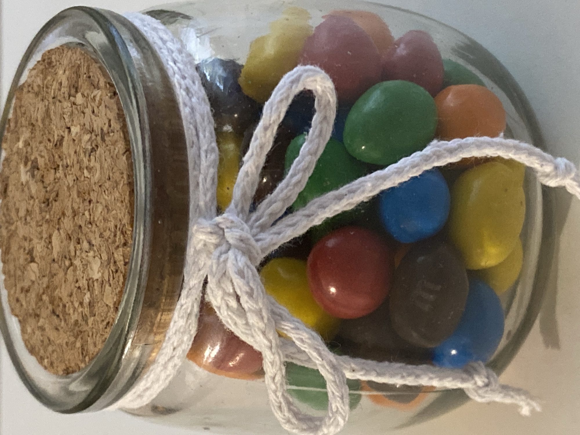 A glass jar filled with colorful candy is sealed with a cork and tied with a white rope. The candy appears to be M&M's or a similar type of candy, with visible colors including red, orange, yellow, green, blue, and brown. The jar is lying on its side, and the rope is tied in a bow around the neck of the jar. The background is a plain white surface. The lighting suggests the photo was taken indoors. There is no visible text in the image.