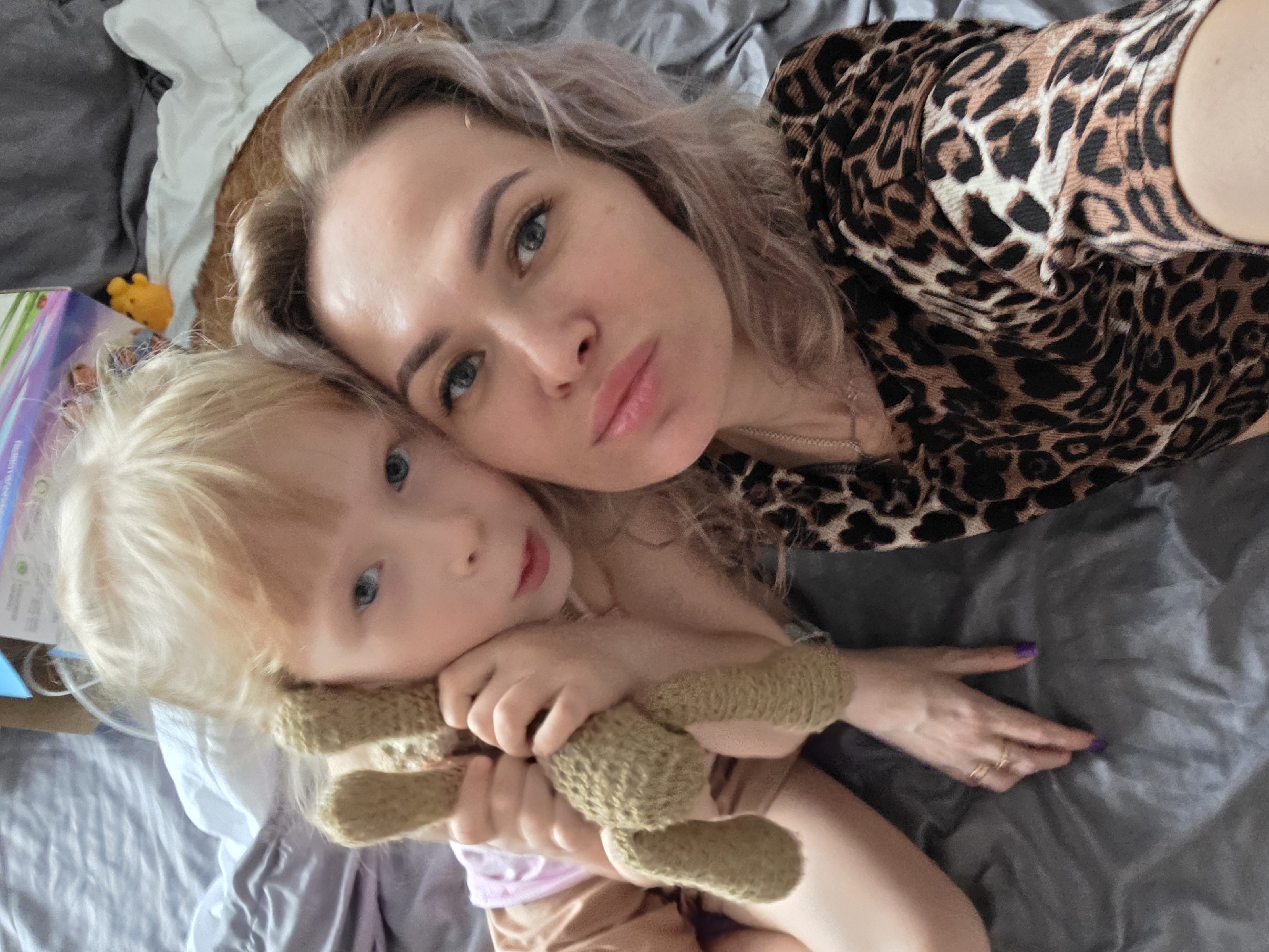 A woman and child are lying on a gray bed. The woman has light hair and is wearing a leopard-print top. The child has blond hair and is holding a stuffed animal. They are both looking at the camera. A box is visible in the background. The location is likely in a residential area, potentially in the city of Chaikovsky, Russia.