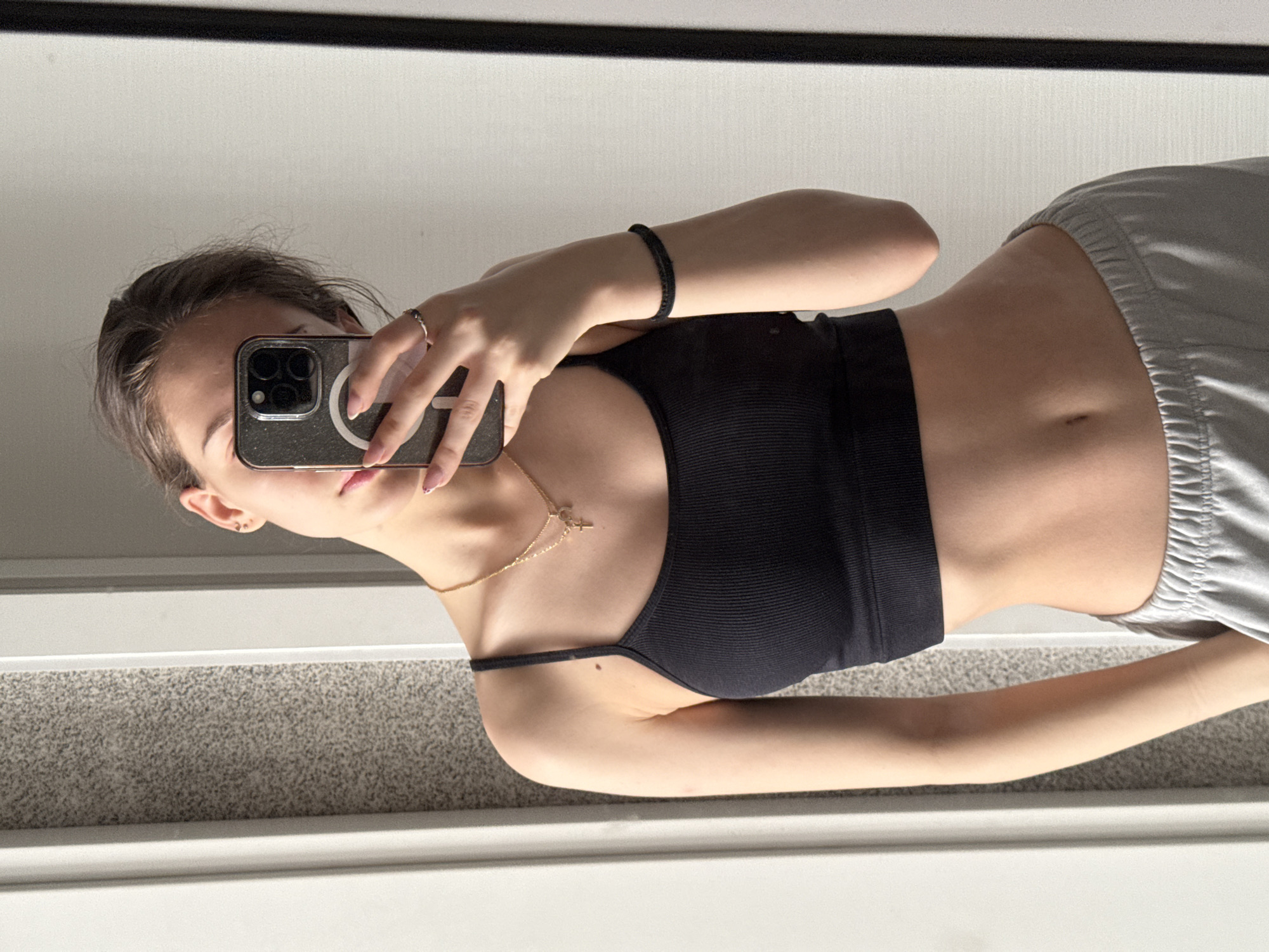 A young woman is taking a selfie in a mirror. She is wearing a black cropped tank top and grey sweatpants. She has a gold necklace and a black bracelet on her wrist. She is holding an iPhone. The background is a white wall, and the floor is carpeted. The location is not discernible as Sofia, Bulgaria.