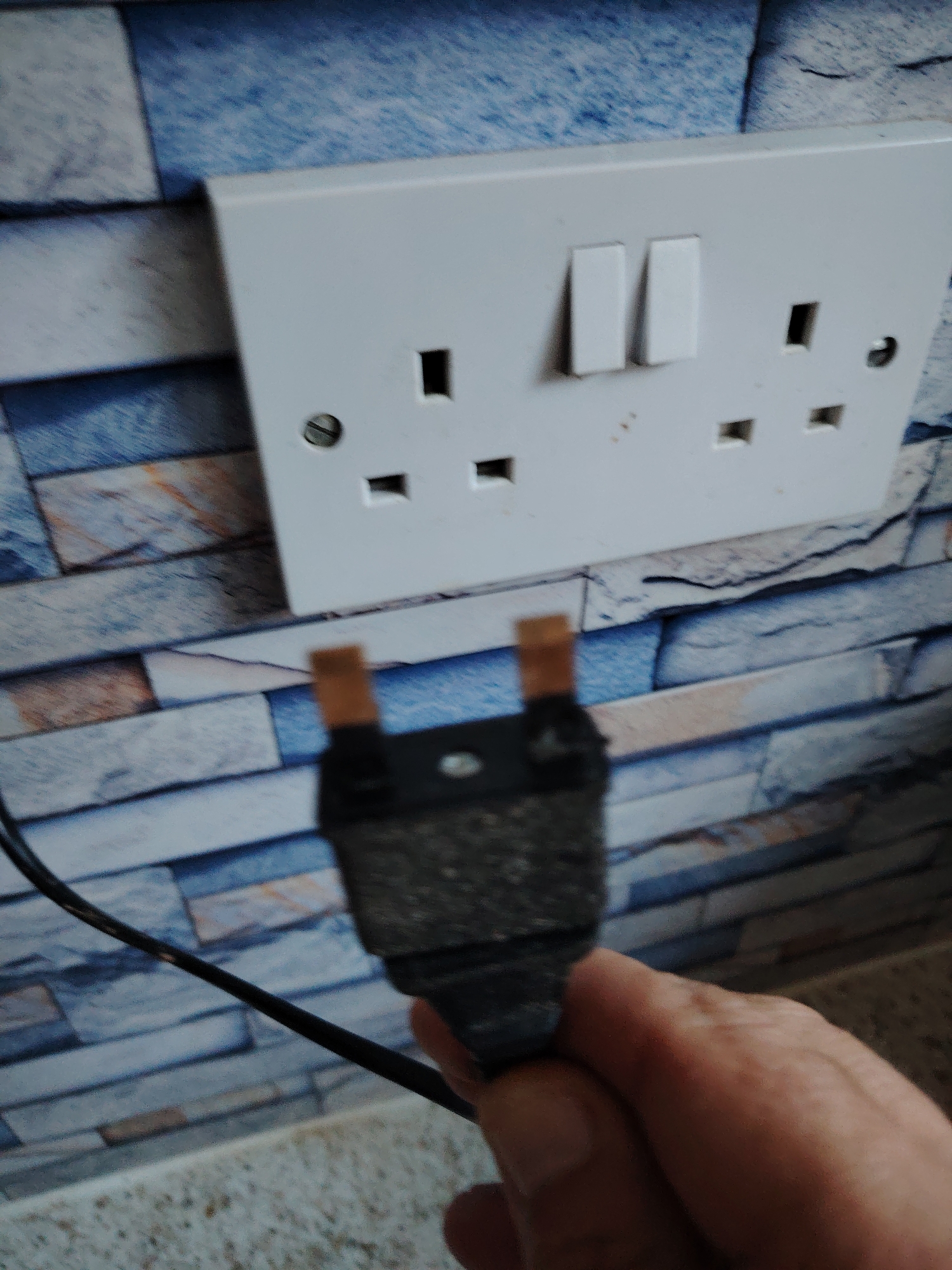 The image shows a close-up of a person's hand holding a black electrical plug with visible metallic prongs against a backdrop of a white double electrical socket mounted on a wall with a blue and white patterned tile effect. The socket has two rectangular switches above the sockets. The scene appears to be indoors. The location in the city of Huntingdonshire, United Kingdom, is not discernible from the image.