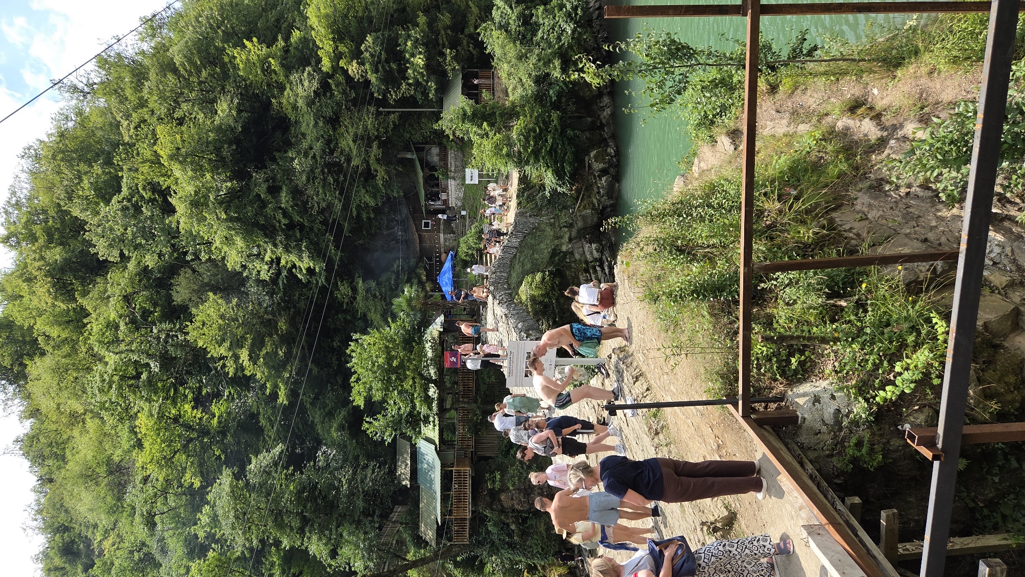 In Kveda Makhuntseti, Georgia, a group of tourists gathers near a historic stone bridge spanning a turquoise river. The bridge, adorned with greenery, leads to a cluster of wooden buildings nestled into a lush, tree-covered hillside. Several people are observed on the bridge and along the riverbank, some appearing to be preparing for or recovering from swimming. The atmosphere is lively, with individuals dressed in casual summer attire. The surrounding environment is characterized by dense foliage and a clear, vibrant body of water.