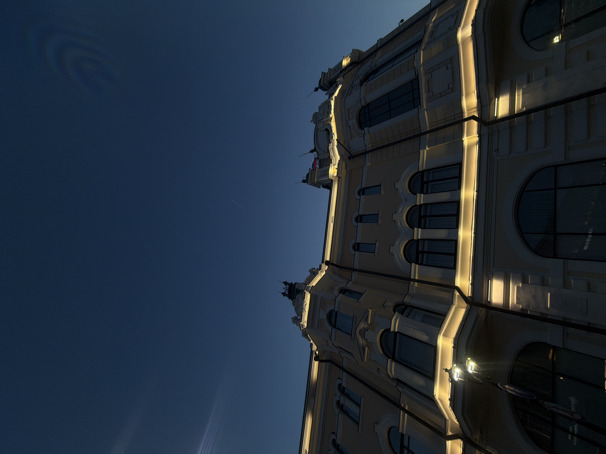 A low-angle shot showcases a building facade in Nizhny Novgorod, Russia, under a clear, dark blue sky. The building is light beige in color and features multiple windows, archways, and decorative architectural elements along its facade. The right side of the building is brightly illuminated with what appears to be artificial lighting, casting a warm glow. A street lamp is visible at the bottom right corner. The sky is mostly empty except for a few light artifacts and what appears to be a faint contrail from an aircraft. The building is angled from the viewer's perspective, emphasizing its height.