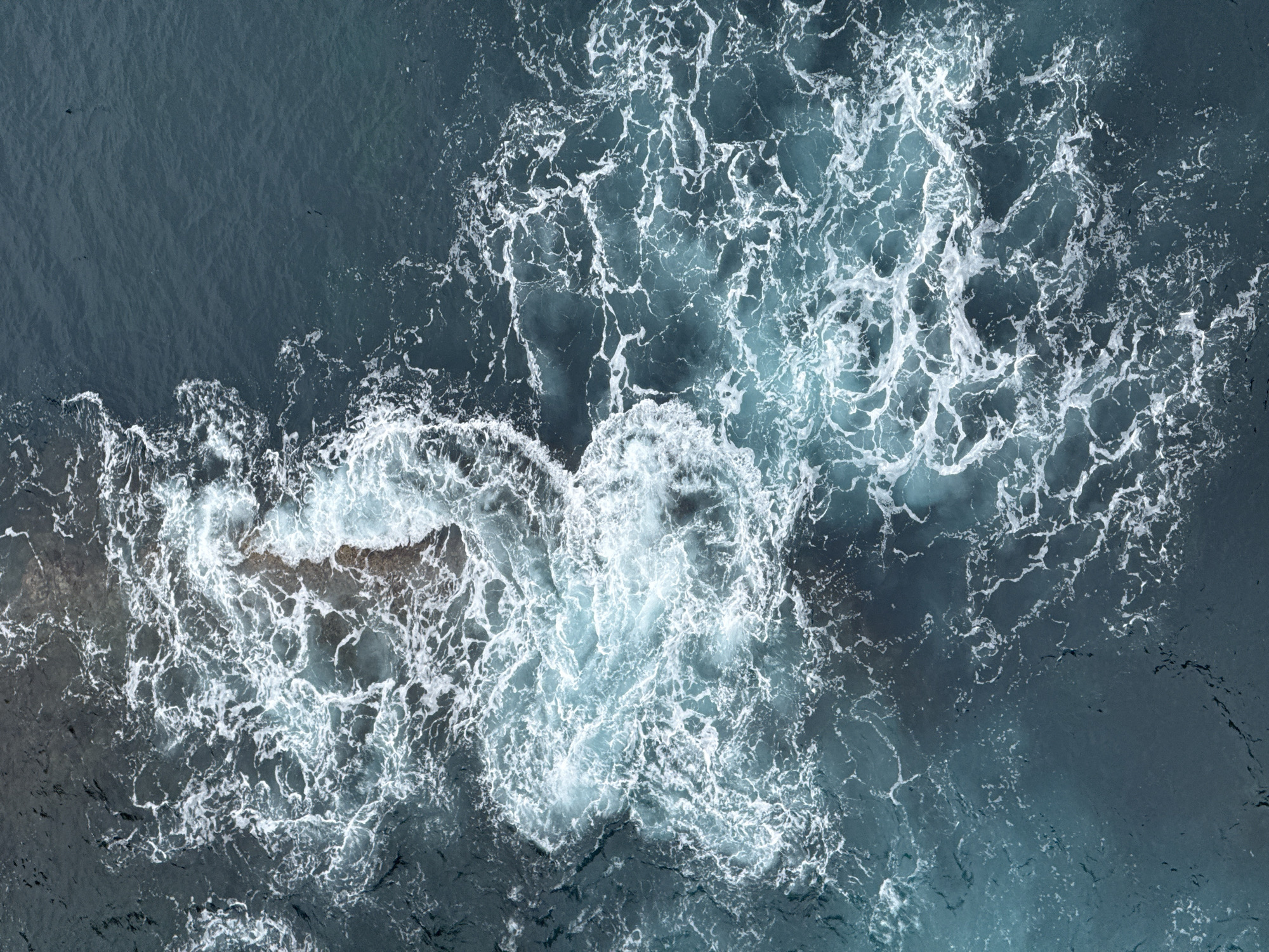 This is an aerial, top-down shot of dark, churning ocean water near a rocky coastline. The water is a deep, dark blue-grey, with areas of lighter, turquoise blue where the waves are breaking and creating foam. White foam and spray are visible as the waves crash against the rocks and churn in the water. The rocky shore appears dark and wet, with some lighter grey and brown tones. The texture of the water is dynamic, with swirling patterns of white foam over the darker depths. There is no visible text. The image appears to be taken during daylight hours, possibly overcast given the muted lighting. The scene evokes a sense of powerful natural forces at work, with the raw energy of the ocean against the rugged coastline. The location cues are consistent with a coastal area, specifically mentioning Cudillero, Spain.