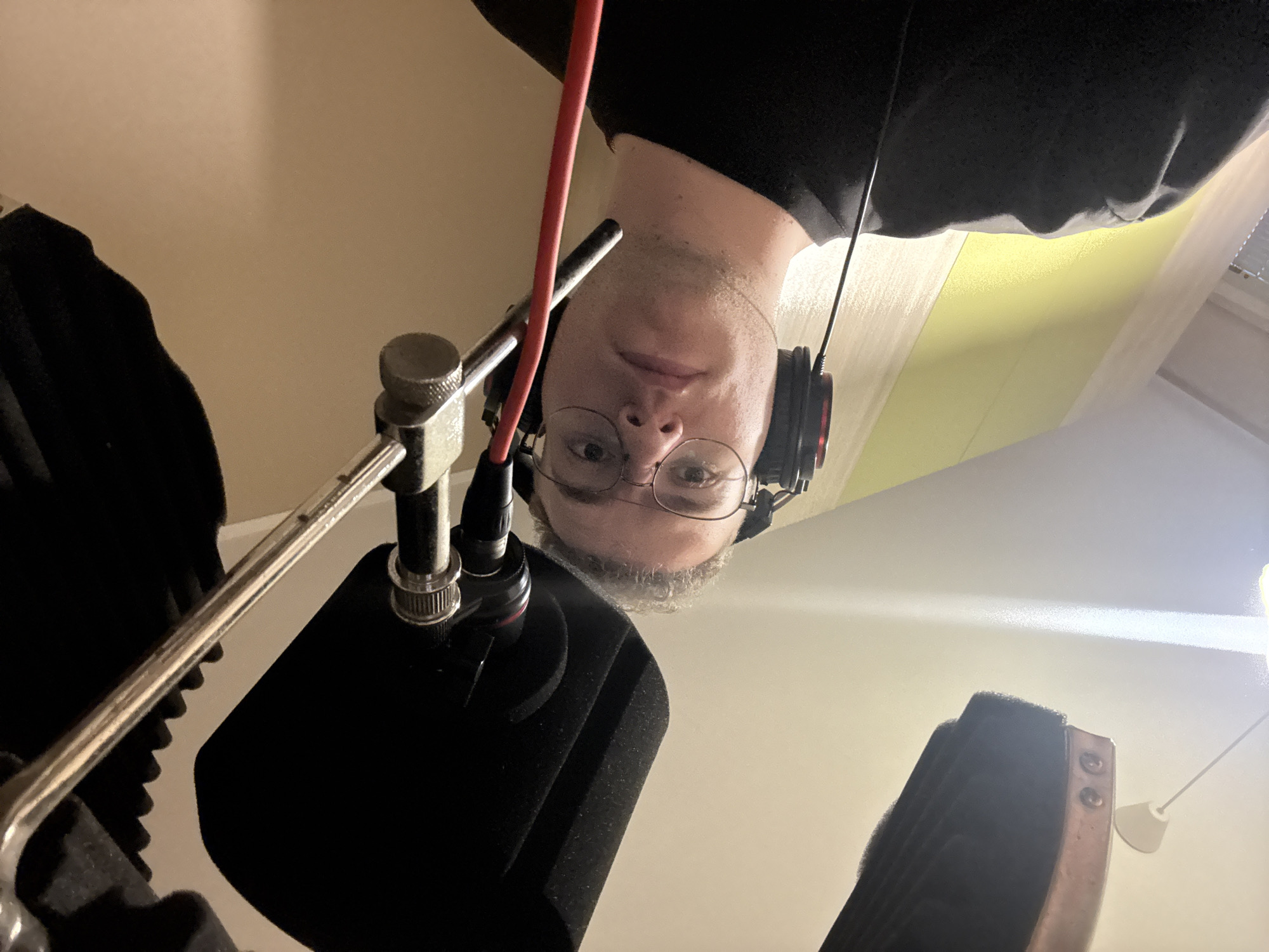 An upward-facing, low-angle shot captures a male individual upside down, positioned over a microphone and recording equipment in an indoor setting, possibly in Plovdiv, Bulgaria. The man, seen from the chest up, wears a dark t-shirt, round glasses, and over-ear headphones. He has short hair and a neutral expression, looking directly at the camera. A prominent red cable extends from near the microphone, running upwards.
The recording setup includes a silver microphone stand holding a black microphone, which is covered by a black foam pop filter. To the left, black acoustic foam panels are partially visible, suggesting a sound-treated environment. The background consists primarily of light beige or cream-colored walls or ceiling, with a bright light source on the upper right, from which a white pull cord hangs. Another section of the background shows a lighter, possibly yellow or green, panel and what appears to be blinds or a window at the top right.
The image suggests the person is engaged in a recording activity such as singing, voice-over work, or podcasting. The unusual perspective implies the photo was taken from below the microphone, possibly as a selfie or by someone else capturing the moment from that angle. No visible text is present in the image.