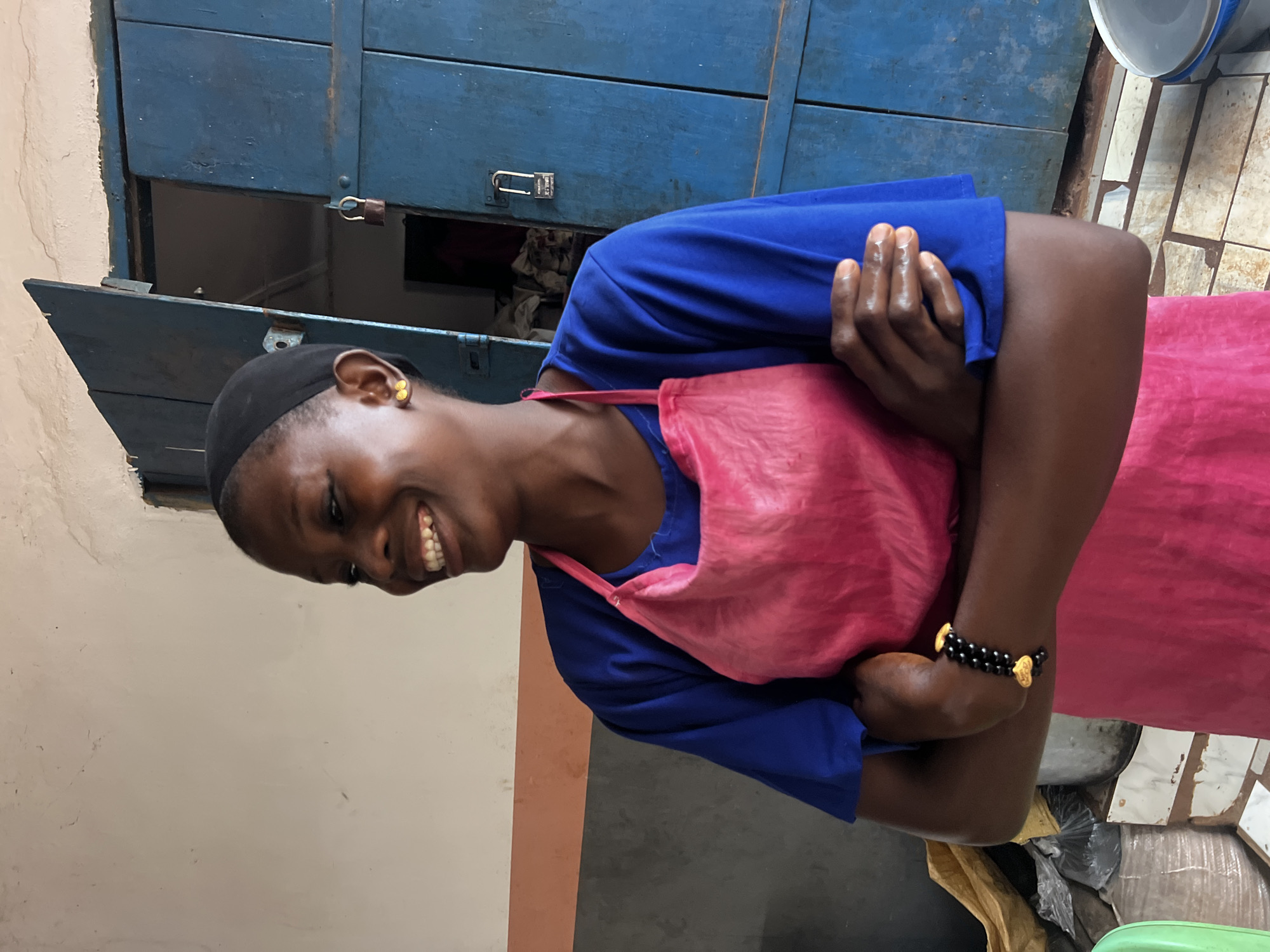 A Black woman is pictured in Nkpor, Nigeria, wearing a blue t-shirt and a bright pink apron. She has her arms crossed and is smiling while looking off to the left. She is wearing gold earrings and a black beaded bracelet with gold heart-shaped charms. Behind her, a large blue wooden door is slightly ajar, revealing a dark interior. The door has two padlocks attached to it. The walls are painted in muted tones of cream and orange. In the foreground, to the left, a portion of a green container is visible, and to the right, a grey bucket sits on the tiled floor.