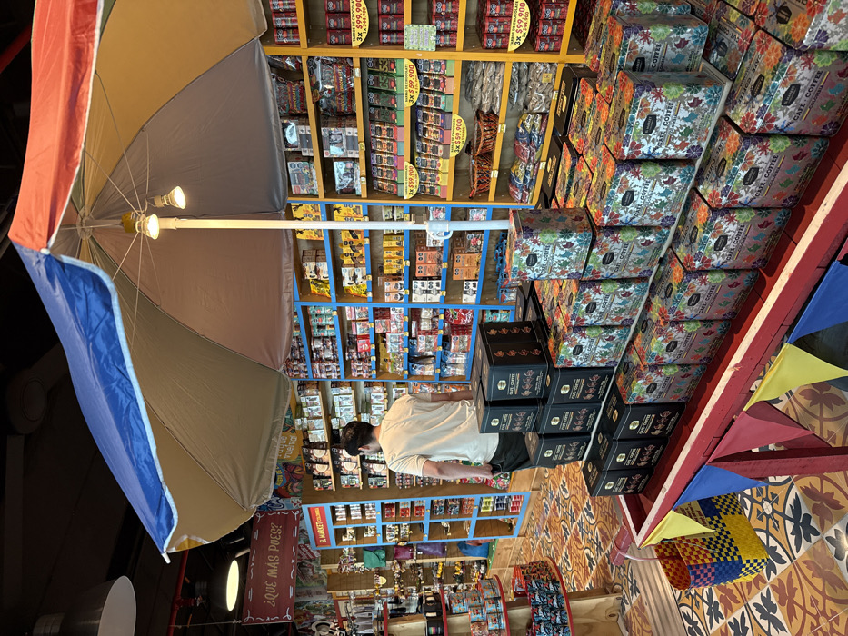 An indoor market scene in the Area Metropolitana Centro Occidente (AMCO), Colombia, is observed from a slightly elevated perspective. A single male individual, seen from behind, with dark hair, wearing a light tan t-shirt and dark pants, stands facing a wall of merchandise shelves on the left side of the frame. In the center foreground, a large, open market umbrella features blue, beige, red, and yellow colored sections, with two small lights attached to its central pole. Beneath the umbrella, a red, boat-shaped display stand, adorned with small red, yellow, and blue pennants, exhibits multiple stacks of rectangular "CAFE COFFEE" tins. Some tins are solid black with gold lettering, while others display a vibrant, multicolored, leaf-like pattern.
Behind the umbrella and the individual, extensive shelving units, colored in bright blue and yellow, line the walls, filled with a diverse array of packaged food products, including coffee, snacks, and numerous chocolate bars with visible pricing, such as "3X $59.900" and "3X $99.900 / 1X $44.900." A hanging wooden sign on the left reads "¿QUÉ MÁS PUES?" and another, "EL MARKET COLOMBIA." The floor features an intricate, multicolored geometric tile pattern. A woven, multicolored basket rests on the floor to the left of the boat display. Overhead, exposed ceiling structures and recessed lighting fixtures are visible.