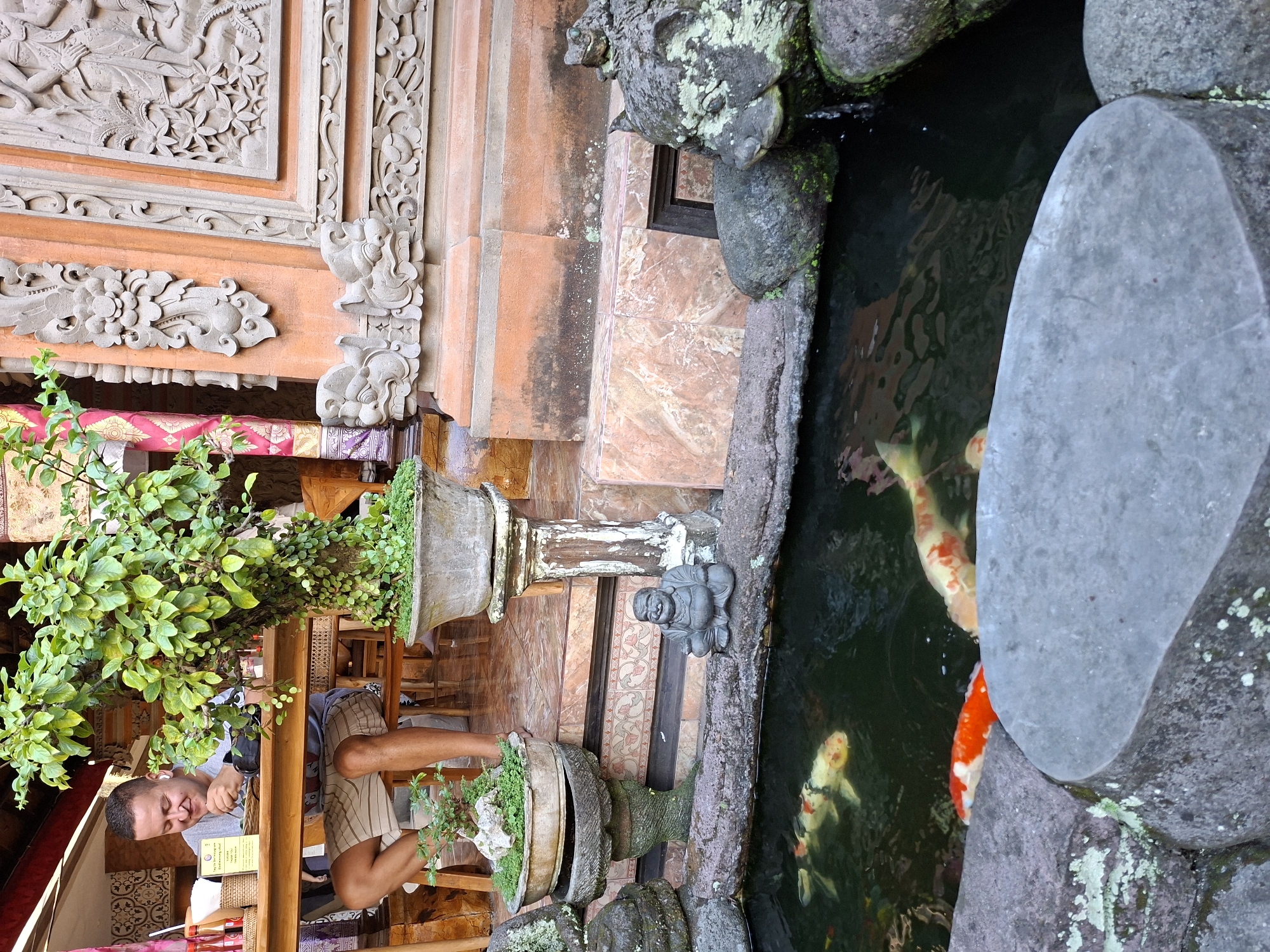 A man sits near a pond at a cafe in the Ubud District of Indonesia. The man is wearing a grey shirt and striped shorts. He sits with one leg crossed over the other and an unidentifiable object is in his hand. The cafe's decor is Balinese. Intricate carvings adorn the walls. Potted plants are placed around the seating area, and the pond contains several koi fish. The pond is surrounded by stones, some covered in moss. A small Buddha statue sits on the edge of the pond. Stairs and tiled floors are visible in the background.