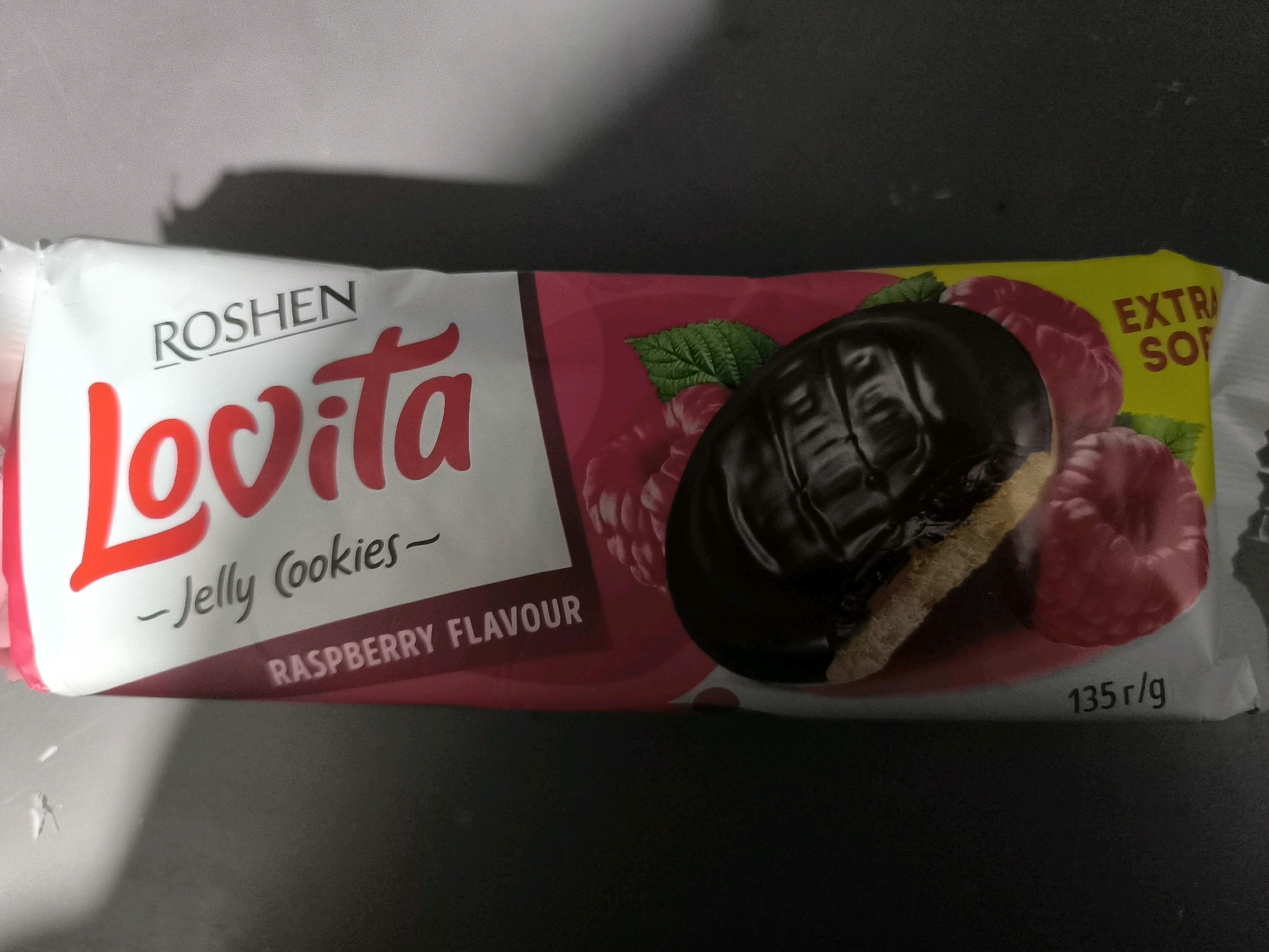 A close-up, overhead shot of a wrapper for "LOVITA" raspberry-flavored jelly cookies is displayed on a dark, reflective surface. The wrapper is predominantly white with a pink and yellow design featuring images of raspberries and a chocolate-covered cookie with visible jelly filling. The brand name "ROSHEN" is printed in black above "LOVITA," which is in large, red, cursive lettering. Below "LOVITA," it reads "-Jelly Cookies-" and "RASPBERRY FLAVOUR" in smaller text. The weight "135 r/g" is visible in the lower right corner. The image is sharp and well-lit, capturing the details of the product packaging. There are no people or any indications of the specific location within Namdanak, Uzbekistan.