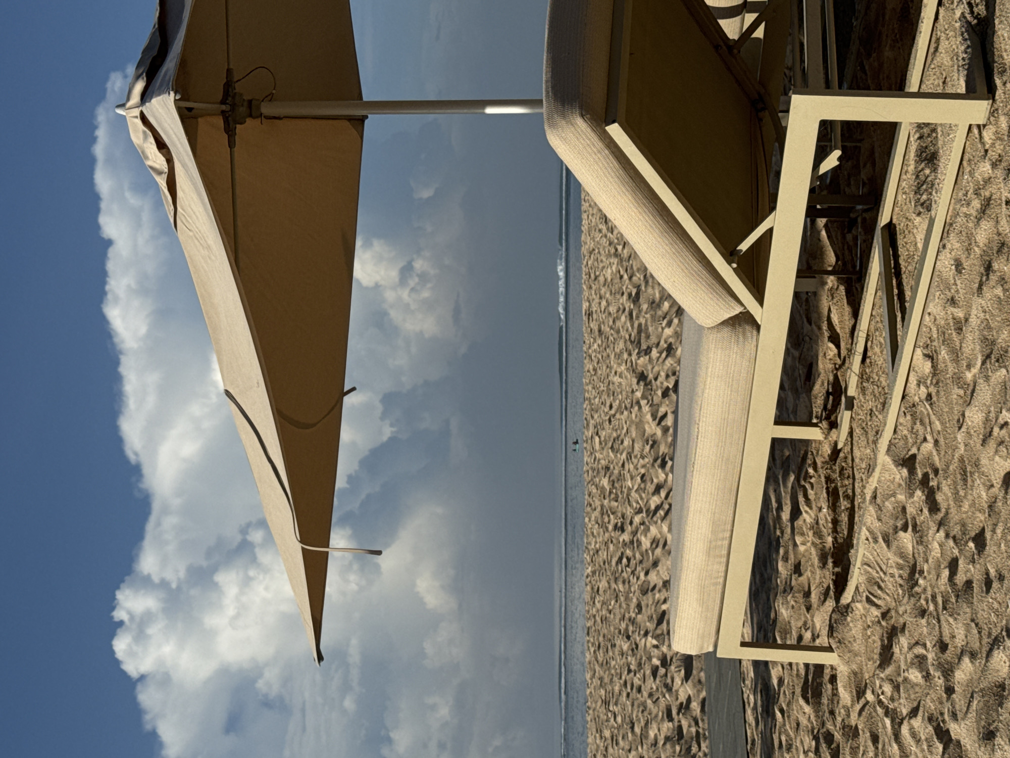 The image depicts a beach scene under a partially cloudy sky, likely in the city of Unknown, Indonesia. A beach umbrella and a lounge chair are prominent in the foreground. The umbrella, with its beige canopy, is tilted towards the left, positioned against a sky featuring alternating blue and cloud-covered areas. The lounge chair, also beige, is oriented towards the right, resting on a sandy beach. The beach stretches towards the horizon, meeting the ocean water under the cloudy sky. The sandy beach appears to have a rough texture, with patterns formed by the wind or the movement of the tide. The scene suggests a serene beach setting, possibly for relaxation and leisure.