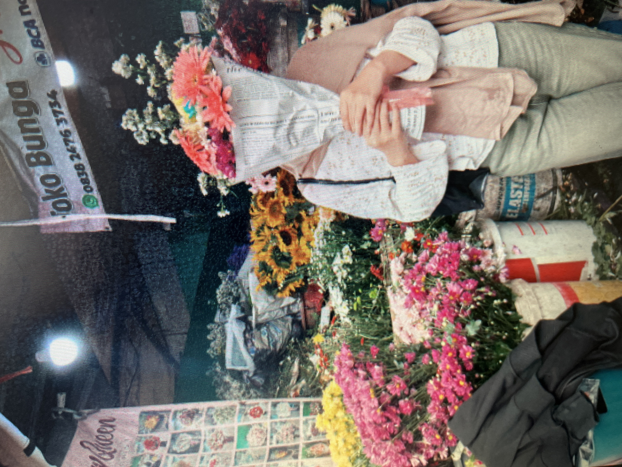 A person stands behind a large bouquet of flowers, their face obscured by the flowers and the white paper wrapping them. They are wearing a white, patterned long-sleeved shirt and light-colored pants. A light brown scarf or shawl is draped over their shoulders and arms. The bouquet is held with both hands, and a pink ribbon is tied around it.
The scene appears to be an outdoor flower stall, likely in Sejahtera, Indonesia. The background is filled with various colorful flowers, including sunflowers, pink chrysanthemums, and yellow flowers. Behind the person, a banner reads "Toko Bunga" (Flower Shop) and displays a phone number and a logo that resembles BCA (Bank Central Asia). Several buckets and containers hold the flowers. A dark garment, possibly a jacket, is draped over some containers in the foreground to the left. The lighting suggests it might be evening or a dimly lit covered area.