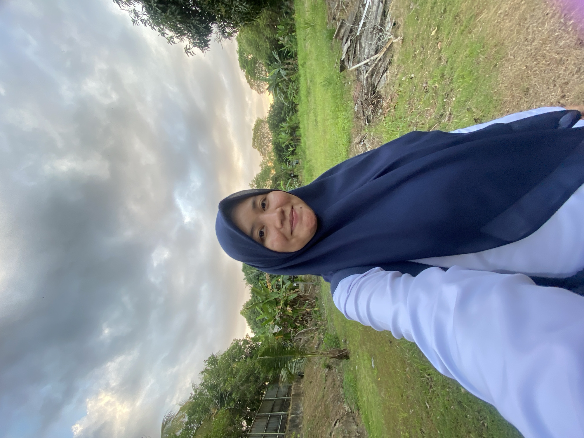 A woman wearing a dark blue hijab and a white long-sleeved shirt is taking a selfie outdoors. She is smiling at the camera. The background is a grassy field with trees and a cloudy sky. The scene appears to be in a rural area.