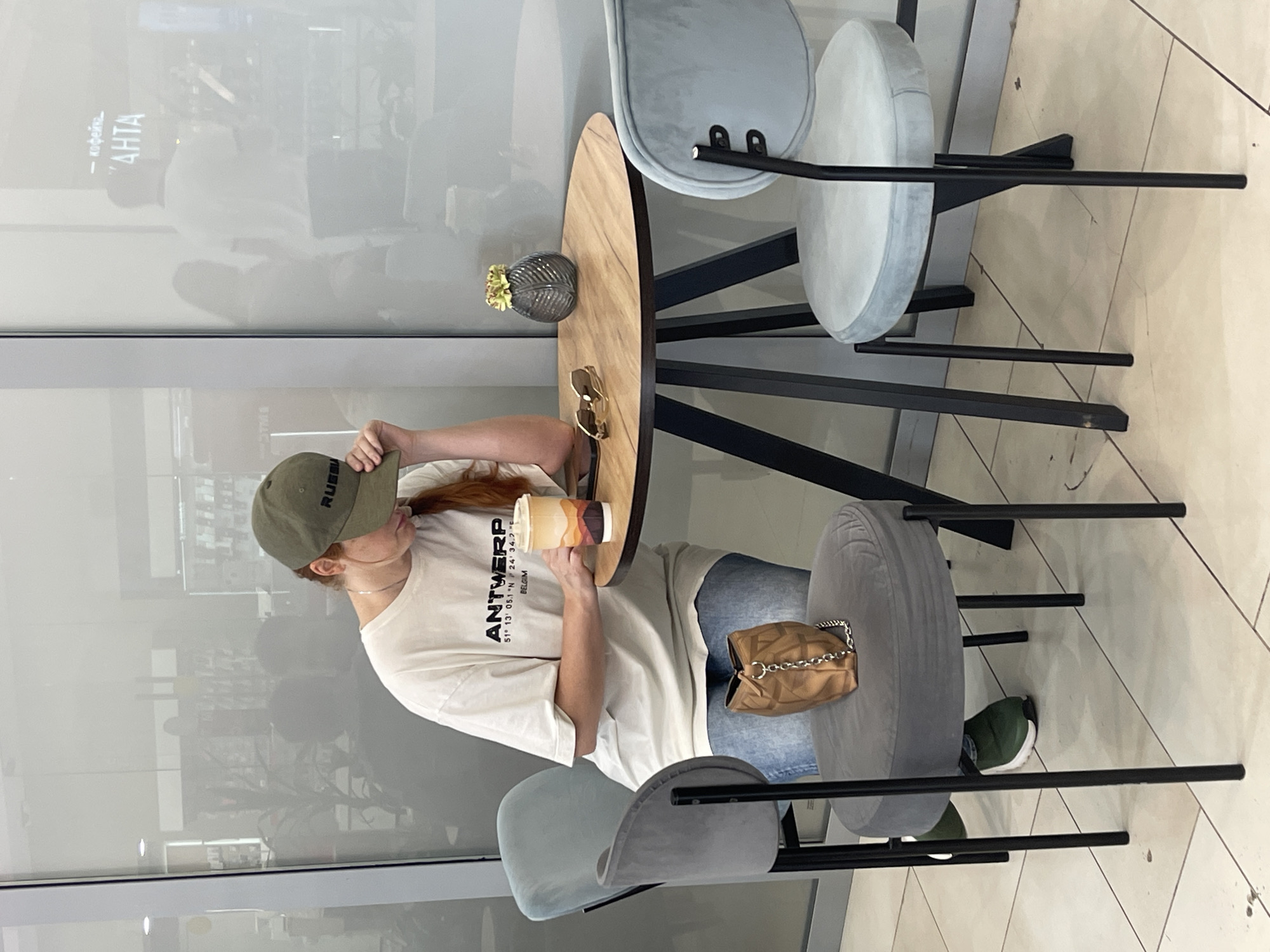 In this photograph, a young woman with red hair is seated at a round wooden table. She is wearing a white t-shirt with the word "ANTWERP" and coordinates printed on it, blue jeans, a green baseball cap, and green sneakers. She holds a takeaway coffee cup in her right hand and her left hand is on the brim of her cap. A small tan purse with a chain strap rests on the seat of a gray chair beside her. The table has a small glass vase with a yellow flower and a pair of sunglasses on it. The scene appears to be indoors, possibly a cafe or restaurant, with large windows reflecting the surroundings. The reflection in the window shows the text "КОФЕЙНЯ" and "АНТА" which translates to "COFFEE HOUSE" and "ANTA". The lighting suggests it could be daytime. The woman's posture and the coffee cup imply she is enjoying a drink. The setting appears modern and casual.