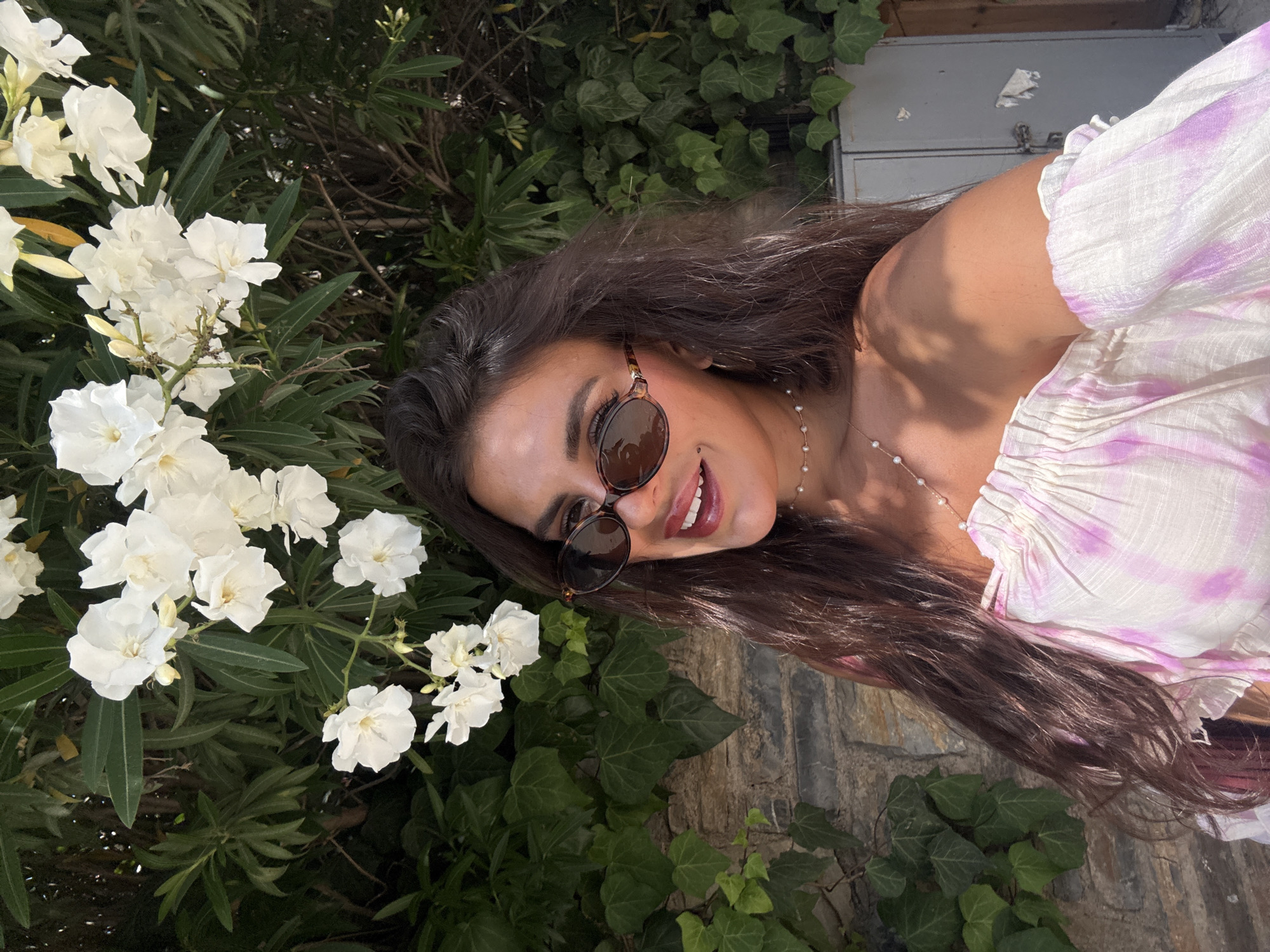 In Bodrum, Turkey, a woman with long, dark hair and sunglasses smiles as she takes a selfie in front of a stone wall overgrown with ivy and a flowering bush with white blossoms. She wears an off-the-shoulder top with a pink and white tie-dye pattern and a delicate pearl necklace. The sun casts a dappled shadow on her shoulder. In the background, part of a gray metal cabinet is visible.