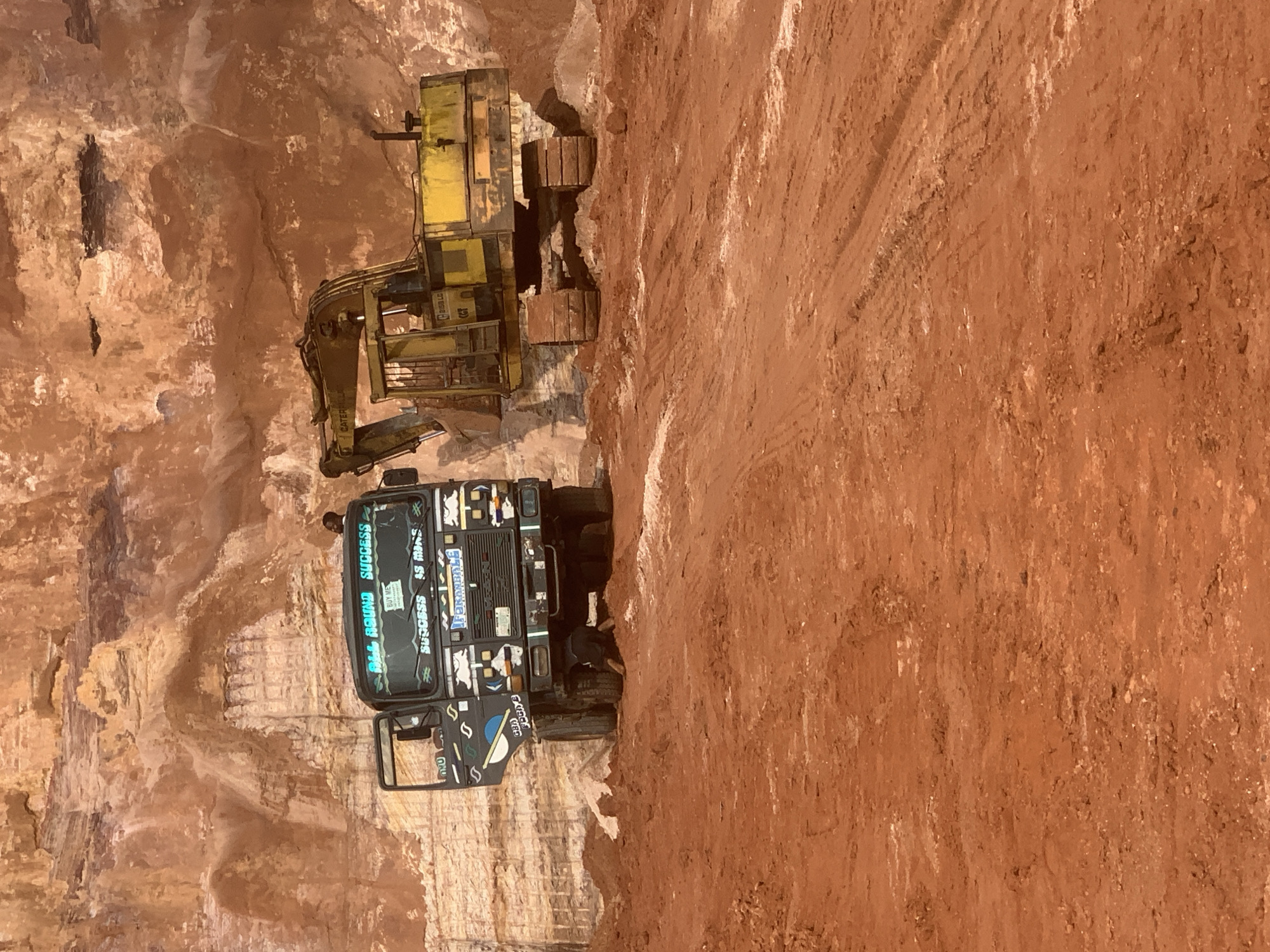 The image depicts a busy construction or mining site in Ago-Iwoye, Nigeria. In the foreground, a red dirt road with tire tracks suggests recent activity. Two large vehicles are present: a dark-colored truck with its door open and an excavator. The truck is adorned with various stickers and inscriptions, including "ALL ROUND SUCCESS" in bright blue lettering on its windshield. A person is partially visible beneath the truck, appearing to be working on it. Behind the truck, a yellow excavator with tracks is positioned, facing towards the right. The backdrop of the scene is a massive, layered rock face, indicative of a quarry or excavation site. The overall impression is one of ongoing industrial work in a rugged, natural setting.