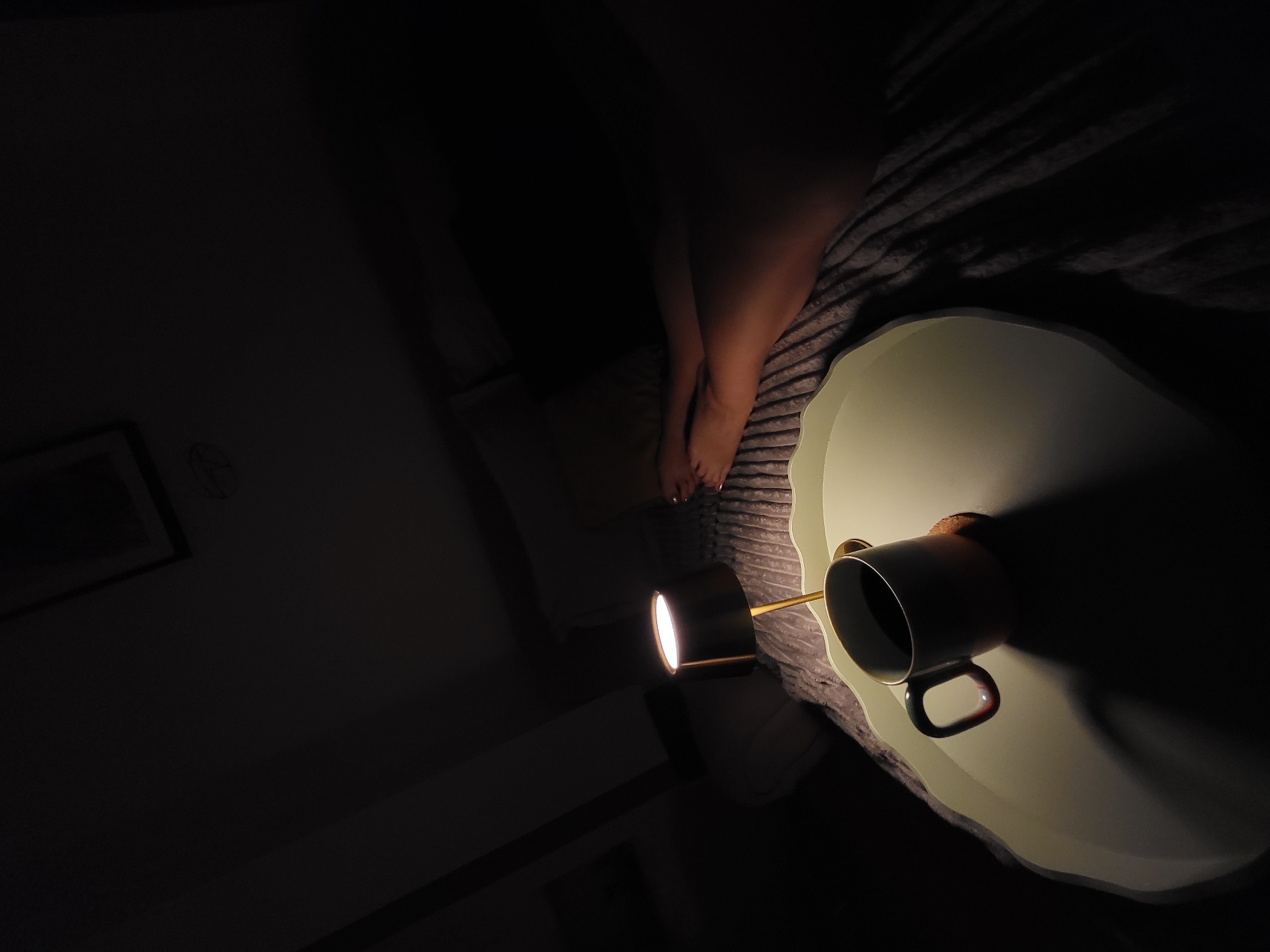 A high-angle shot in a dimly lit room features a cup resting on a decorative plate or tray. The cup, dark in color with a handle, casts a significant shadow across the plate. A light source, possibly a small lamp, is positioned near the cup, illuminating the plate. The background is dark, but one can discern a bed and a pair of legs. A picture hangs on the wall in the background. The location (Budva, Montenegro) cannot be verified from the image.