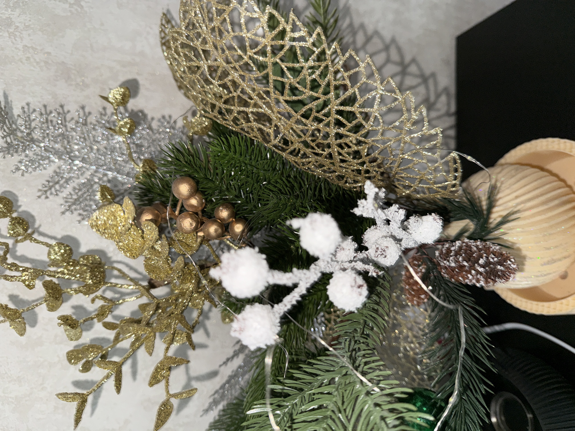 This image depicts a close-up, still-life arrangement of artificial Christmas decorations. The arrangement features elements such as glitter-covered branches, gold berries, white frosted berries, pine sprigs, pinecones, and a gold mesh ribbon. The decorations are clustered together and appear to be part of a larger decorative piece or centerpiece. The background is a textured, light-colored wall, and the decorations are placed on a dark surface, possibly a table or shelf. The lighting suggests an indoor setting. There are no people or animals present in the image. The location of Malmyzh, Russia, is not visually indicated in the image itself and would require external information to be associated with this scene.