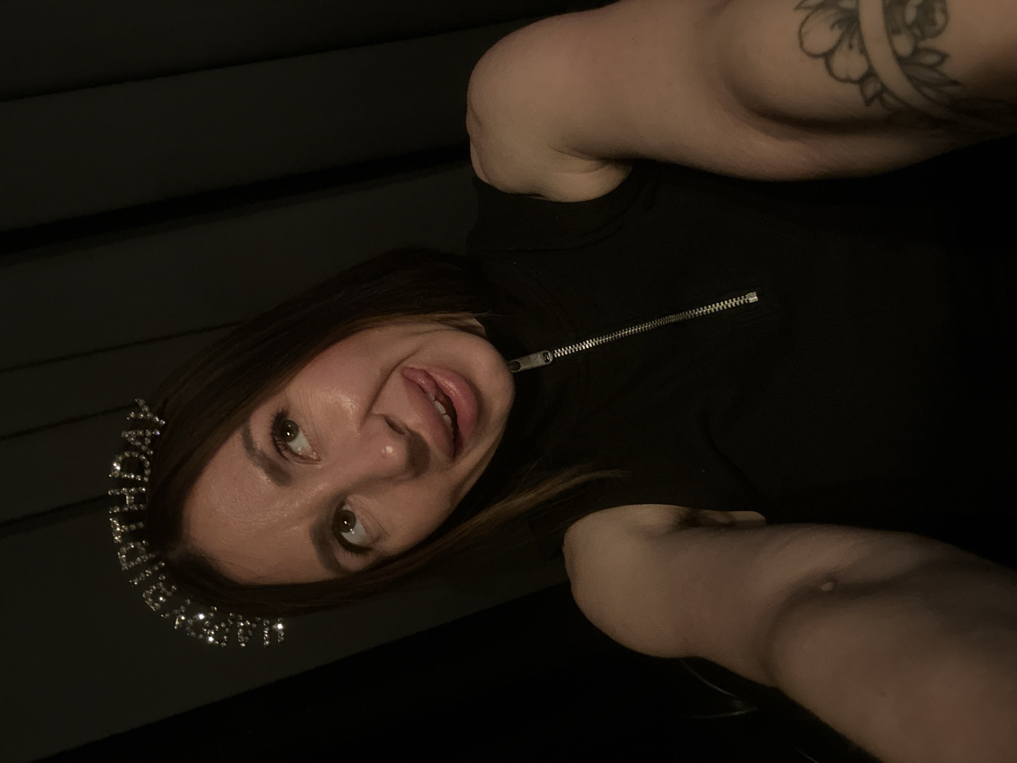 A woman is taking a selfie. She is wearing a black top with a zipper, and a "Happy Birthday" tiara. She has dark brown hair and light skin. She is sticking her tongue out slightly. A tattoo is visible on her upper arm. The background is dark, appearing to be indoors. There is no evidence indicating the location is Votkinsk, Russia.