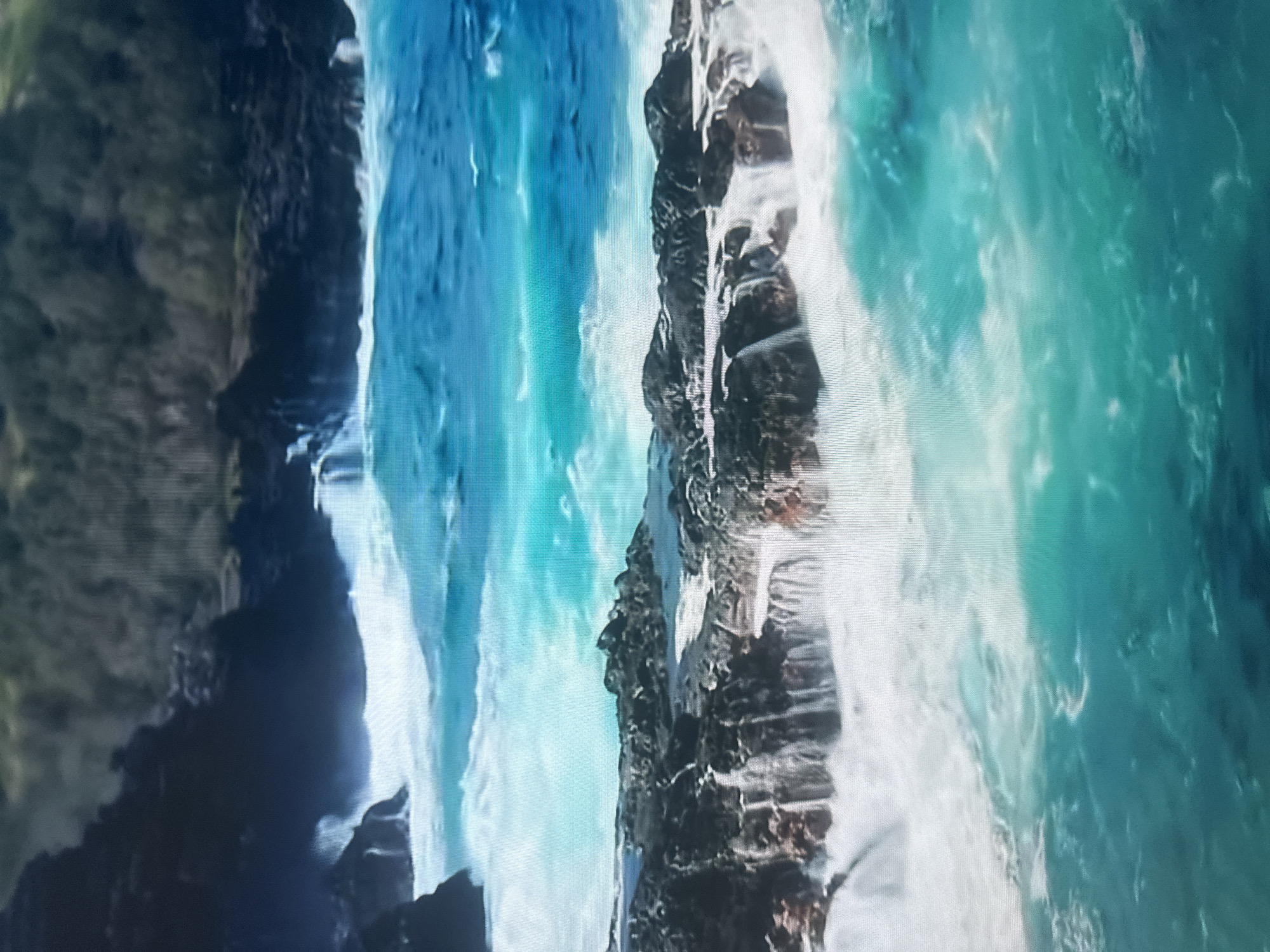 The image depicts a dramatic coastal scene with powerful waves crashing against rocky cliffs. The water is a vibrant turquoise and blue, with white foam and spray indicating the force of the ocean. Jagged, dark rocks form the coastline, with some appearing wet and slick from the impact of the waves. The perspective is from above, looking down at the turbulent water and the rugged terrain. The scene evokes a sense of raw natural power and beauty. There are no people, objects, or visible text in the image. The time of day and weather are not explicitly discernible, but the scene suggests an active, possibly windy, coastal environment. The location context of Almaty, Kazakhstan, is unusual for this type of imagery, as Almaty is a landlocked city.