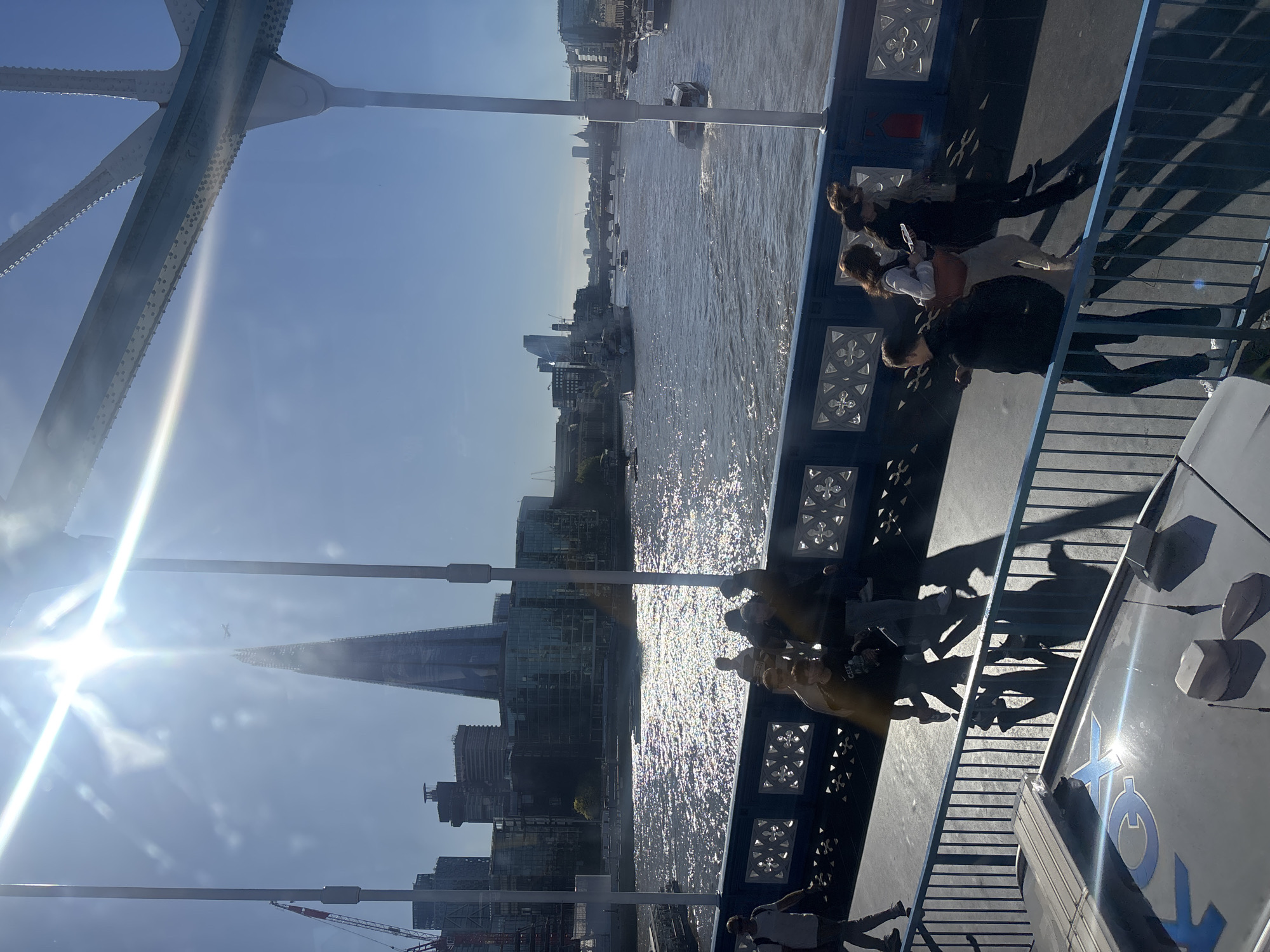 The image captures a vibrant, sunlit scene from what appears to be the pedestrian walkway of Tower Bridge in London, UK, looking southeast along the River Thames. The perspective is slightly rotated clockwise, with the river running from the upper left to the lower right of the frame if viewed horizontally.
In the foreground and to the right, several individuals are seen walking or standing on the bridge walkway. Their elongated shadows are cast dramatically across the path, indicating a bright, sunny day with the sun at a moderate angle. The ornate, dark blue railings with white decorative patterns of Tower Bridge are clearly visible, separating the walkway from the river below.
The River Thames dominates the mid-ground, its surface sparkling intensely with sun reflections, creating a dazzling effect. Several boats, including what appears to be a smaller vessel and a larger boat, navigate the waterway.
On the far bank, a dense urban landscape unfolds, featuring numerous modern buildings and skyscrapers. The most prominent landmark is The Shard, a distinctively tall, triangular glass building, rising majestically into the clear blue sky. Construction cranes are also visible in the distance, suggesting ongoing development in the city.
Part of the intricate metal superstructure of Tower Bridge, with its riveted beams and supporting struts, is visible in the upper left portion of the image. A strong sunburst with lens flare emanates from the upper left, contributing to the bright atmosphere and partially obscuring the view in that area.
The time of day appears to be mid-morning or afternoon, characterized by the strong sunshine and distinct shadows. The weather is clear and bright with no visible clouds. On a grey surface in the bottom right, a partially visible blue logo or text can be seen, which appears to start with "TO", likely part of "TOWER BRIDGE" or a related attraction branding. The overall mood is lively and active, characteristic of a popular tourist destination on a sunny day.