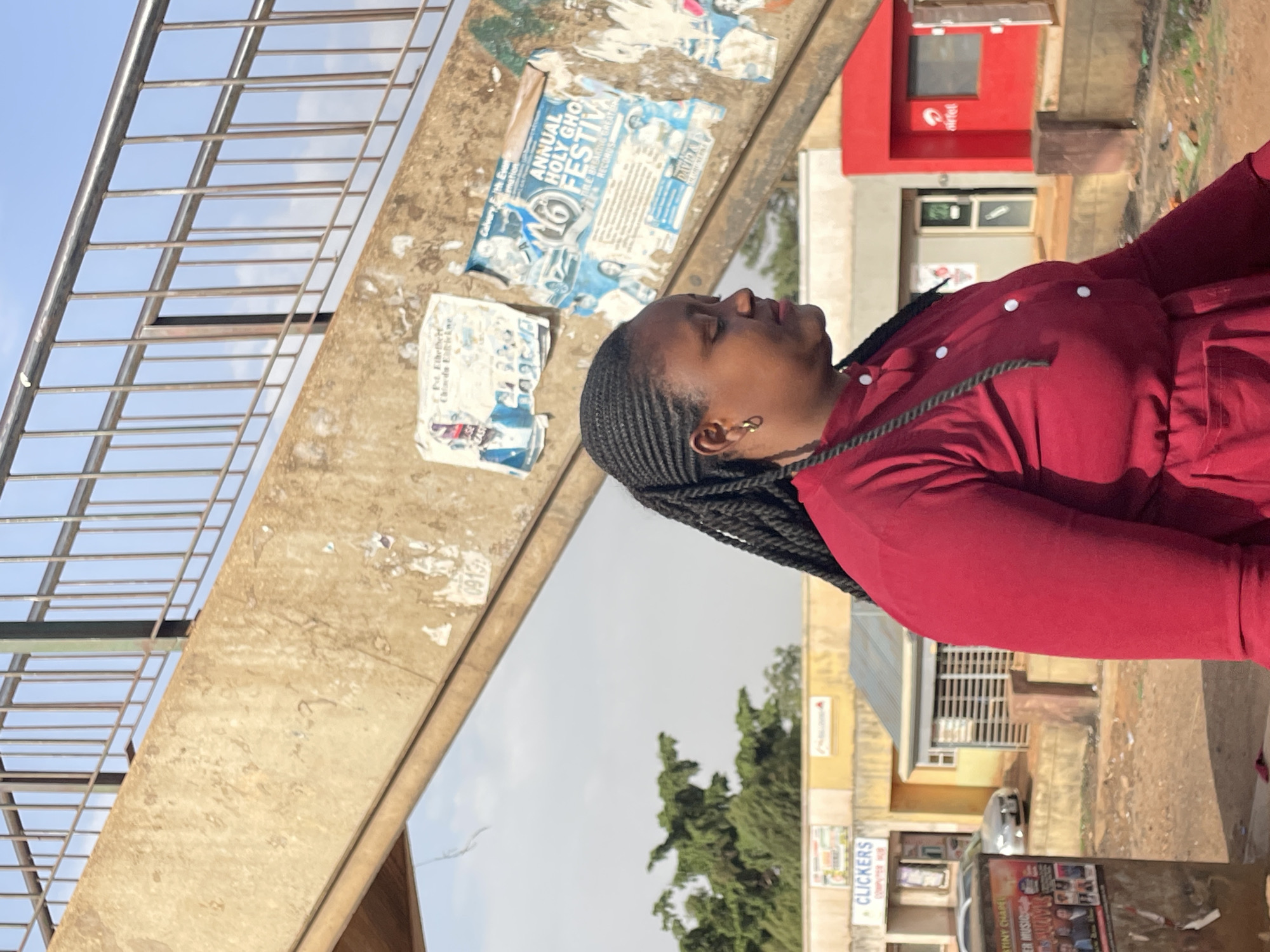 A young woman with braided hair, wearing a red dress, stands on a pedestrian bridge. On the bridge's concrete support are several posters, including one for an "Annual Holy Ghost Festival". An Airtel kiosk is visible in the background. Across the street, a business called "Clickers Computer Hub" is identifiable. The location is Jos, Nigeria.