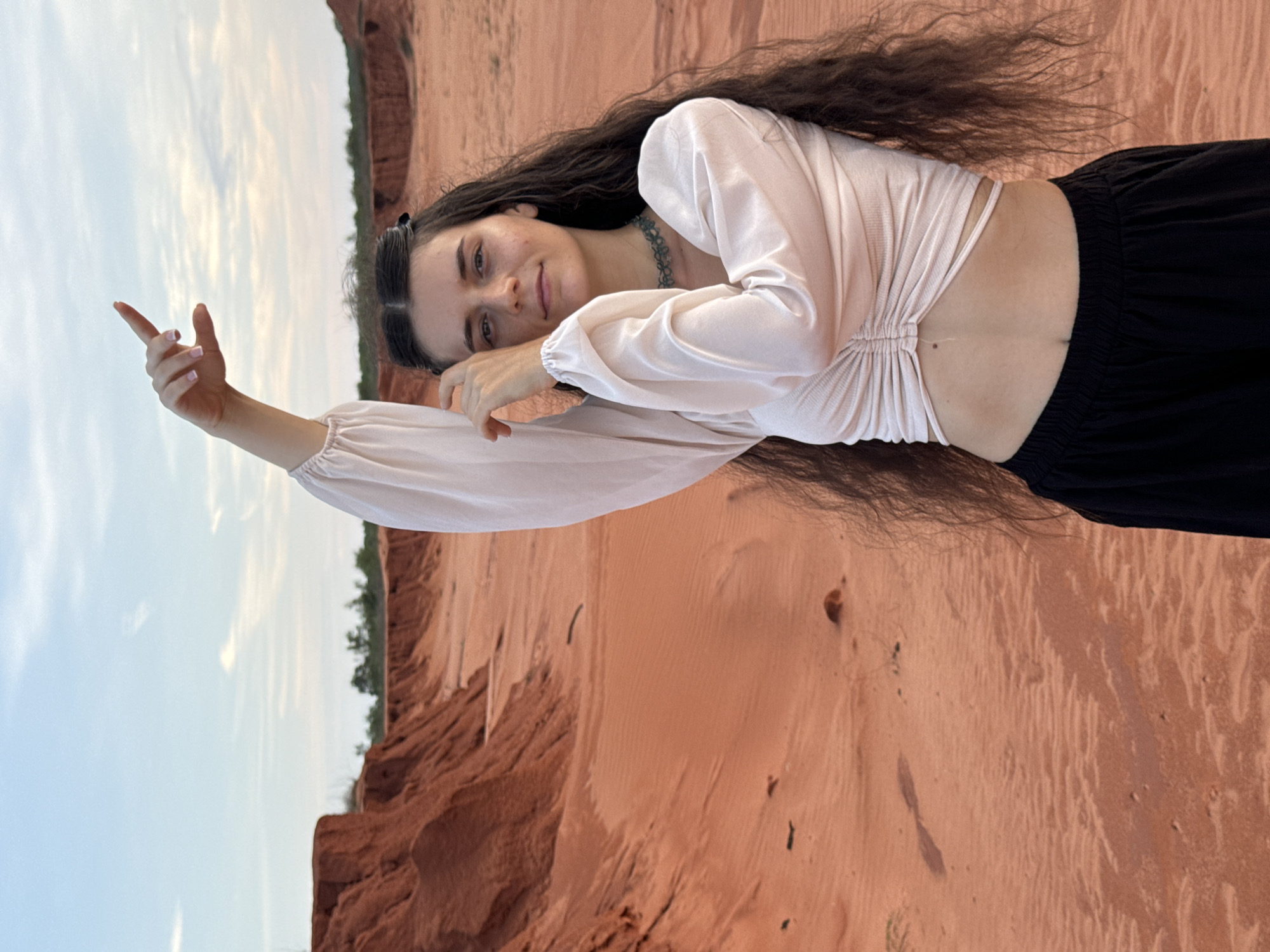 A young woman is depicted lying on her side on reddish-orange sand dunes under a partly cloudy sky. She has long, dark, wavy hair and appears to be in her late teens or early twenties. She is wearing a light-colored, possibly pale pink or cream, long-sleeved crop top with ruched detailing on the front, revealing her midriff. Her lower body is clad in loose-fitting black pants. A dark-colored, possibly beaded or patterned, necklace adorns her neck.
She is looking towards the viewer with a subtle, relaxed smile, her head resting on her left forearm. Her right arm is extended upwards and slightly forward, with her index finger pointing towards the upper left portion of the frame (sky). The sand around her shows distinct ripples and textures, characteristic of wind-blown dunes. In the background, beyond the immediate sandy expanse, there are larger reddish-brown geological formations, resembling cliffs or mounds, topped with sparse green vegetation. The sky is a light blue with scattered white clouds, suggesting it is daytime with soft, diffused light. No text is visible in the image. The scene captures a moment of calm and natural beauty in a desert-like environment, consistent with the sand dunes of Mui Ne, Vietnam.