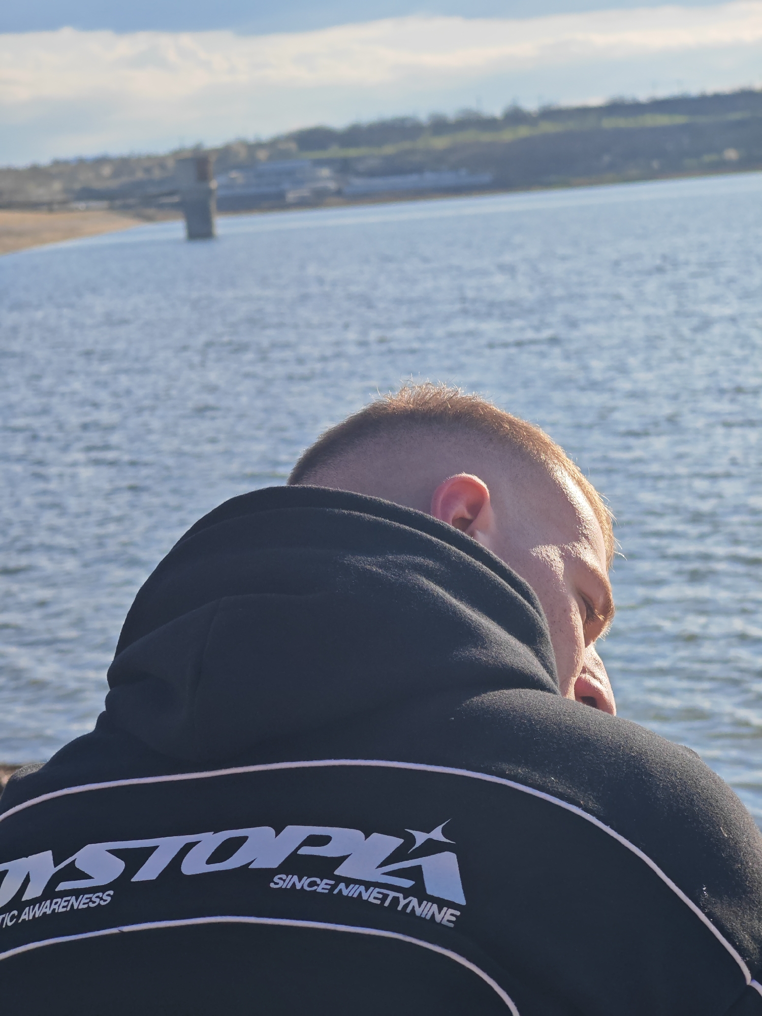 The image shows a man with short, blond hair, wearing a black hooded sweatshirt with white lettering "DYSTOPIA SINCE NINETYNINE" on the back, facing away from the camera. The background consists of a body of water, potentially a lake or reservoir, with a distant structure in the water and the shoreline with some greenery and buildings visible on the horizon.
Given the information that this image relates to the city of Shumen, Bulgaria, the body of water is likely the язовир "Тича" (Ticha Dam Lake), located near Shumen.