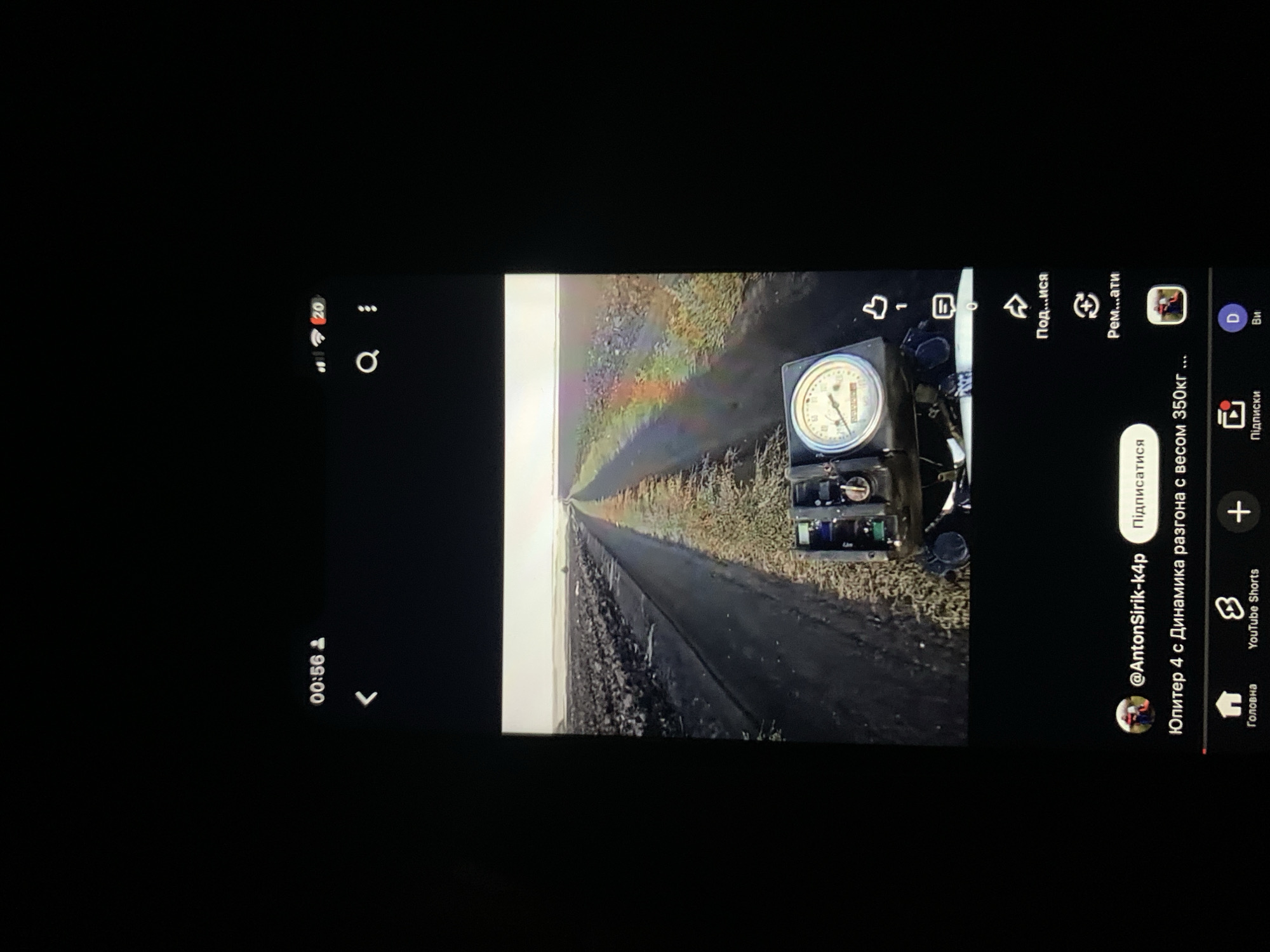 The image shows a screenshot of a YouTube video. The video appears to be a first-person perspective shot from a vehicle, possibly a motorcycle, driving through a field. The field is covered with dark plastic sheeting, and there is a dirt track or road running through it. In the foreground, a gauge or meter is visible, displaying readings. The top of the screen shows a time of 00:56. The bottom of the screen displays YouTube interface elements, including the channel name "@AntonSirik-k4p", the title "Юпитер 4 с Динамика разгона с весом 350кг ...", and navigation icons for "Головна", "YouTube Shorts", "Підписки", and "Ви". There is no discernible information within the image to suggest a specific location in the city of Zhmerynka, Ukraine, or any other geographical context.