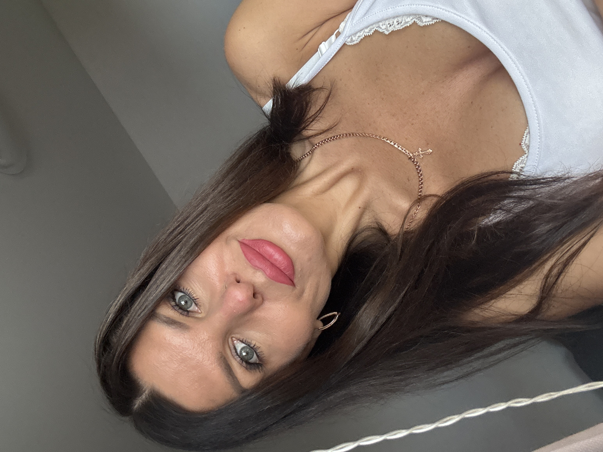 A woman with long, dark brown hair and blue-green eyes is pictured in a selfie. She is wearing a white camisole with lace trim and a rose gold necklace with a small cross pendant. She has on hoop earrings and is wearing pink lipstick. Her head is tilted, and she is looking at the camera. The background is out of focus and appears to be an indoor setting with neutral-colored walls. There are no discernible landmarks or indicators to suggest a specific location in Chaikovsky, Russia.